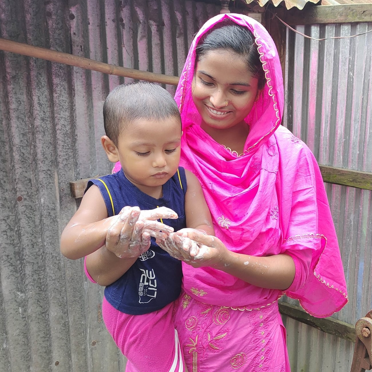 Health leader Sabikunnahar, wearing a bright pink traditional outfit, smiles while helping a young child wash their hands with soap and water outdoors beside a corrugated tin wall.