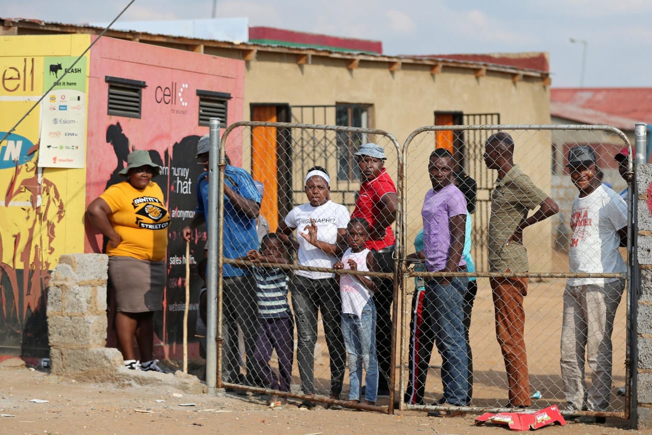 Residents look from behind a closed gate as members of the military patrol during a nationwide lockdown aimed at limiting the spread of novel coronavirus—in Soweto, South Africa on April 23, 2020. The photo shows several people congregated behind a closed fence though there is a very large gap between the fence and the adjacent wall. REUTERS/Siphiwe Sibeko