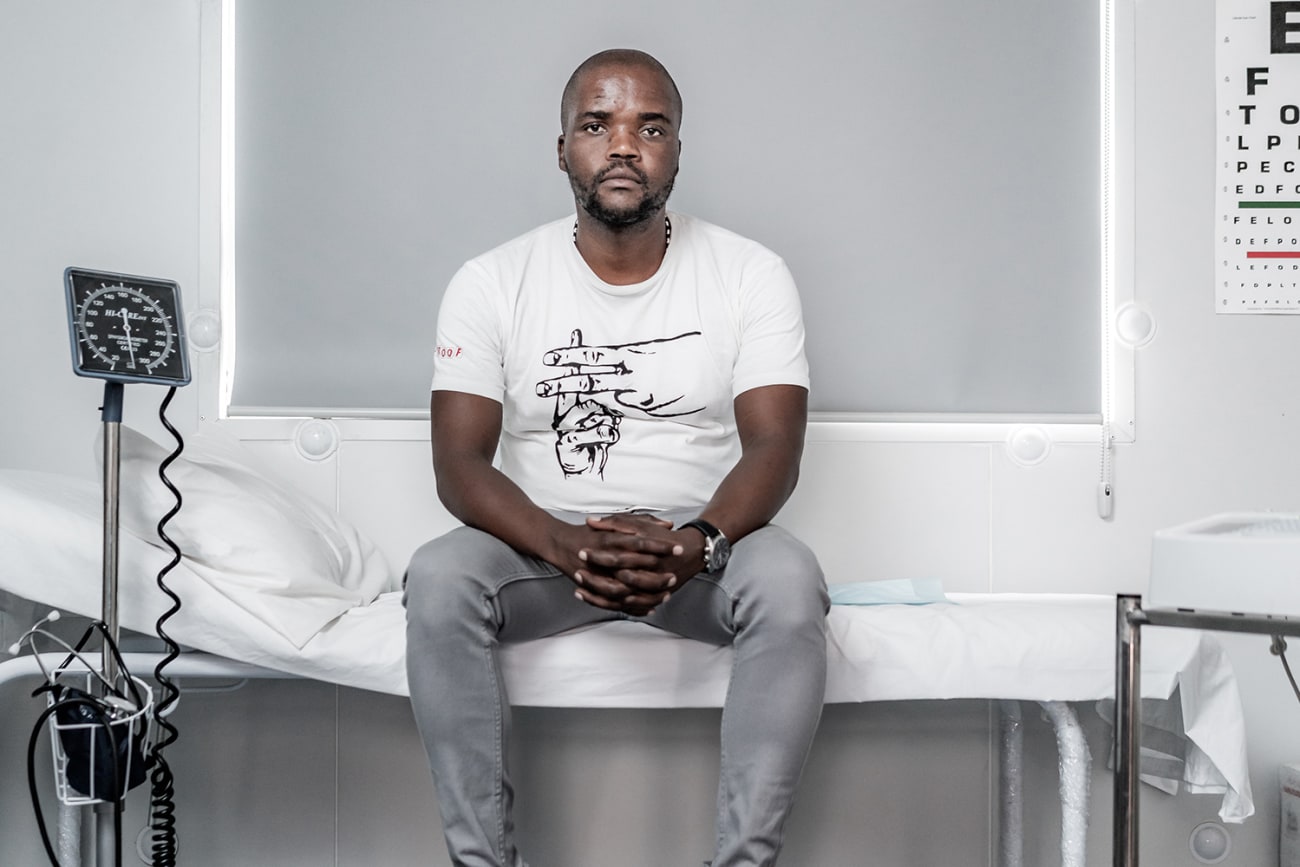 Goodman Makanda, who's involved in efforts to make tuberculosis and COVID-19 testing more acceptable and accessible for South African men, in Khayelitsha, Cape Town, South Africa, on March 24th 2019. The photo shows a man sitting in a clinic with various monitors and equipment. An eye exam is visible on the wall. MSF/Jelle Krings