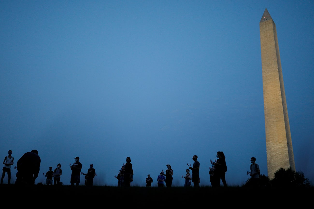 They are informally honored—but are they officially counted? Activists stand at sunrise to memorialize victims of the coronavirus, near the Washington Monument in Washington, DC on August 27, 2020. The photo shows the monument in the thin morning lite with a silhouette line of people standing in the foreground. REUTERS/Tom Brenner