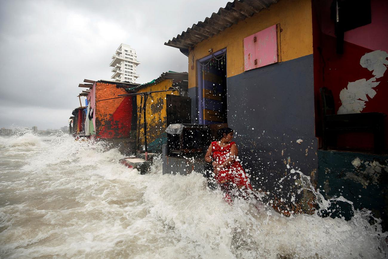 A woman is standing in front of a red, yellow, and grey slate colored tin-roof building with an open door. It is one of several structures in a row—there's an orange one, a green one, and a yellow one. She is turning her head away from the violence of the crashing wave, which sends water shooting up all around her and nearly to the top of the building. All the area in front of the structure is covered by turbulent water. This is a frightening scene.