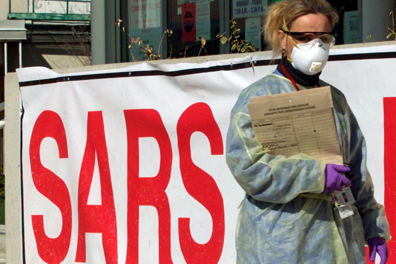 The image shows a woman wearing full protective gear, including eye protection and a surgical mask in front of a hospital adorned with a huge sign that says 