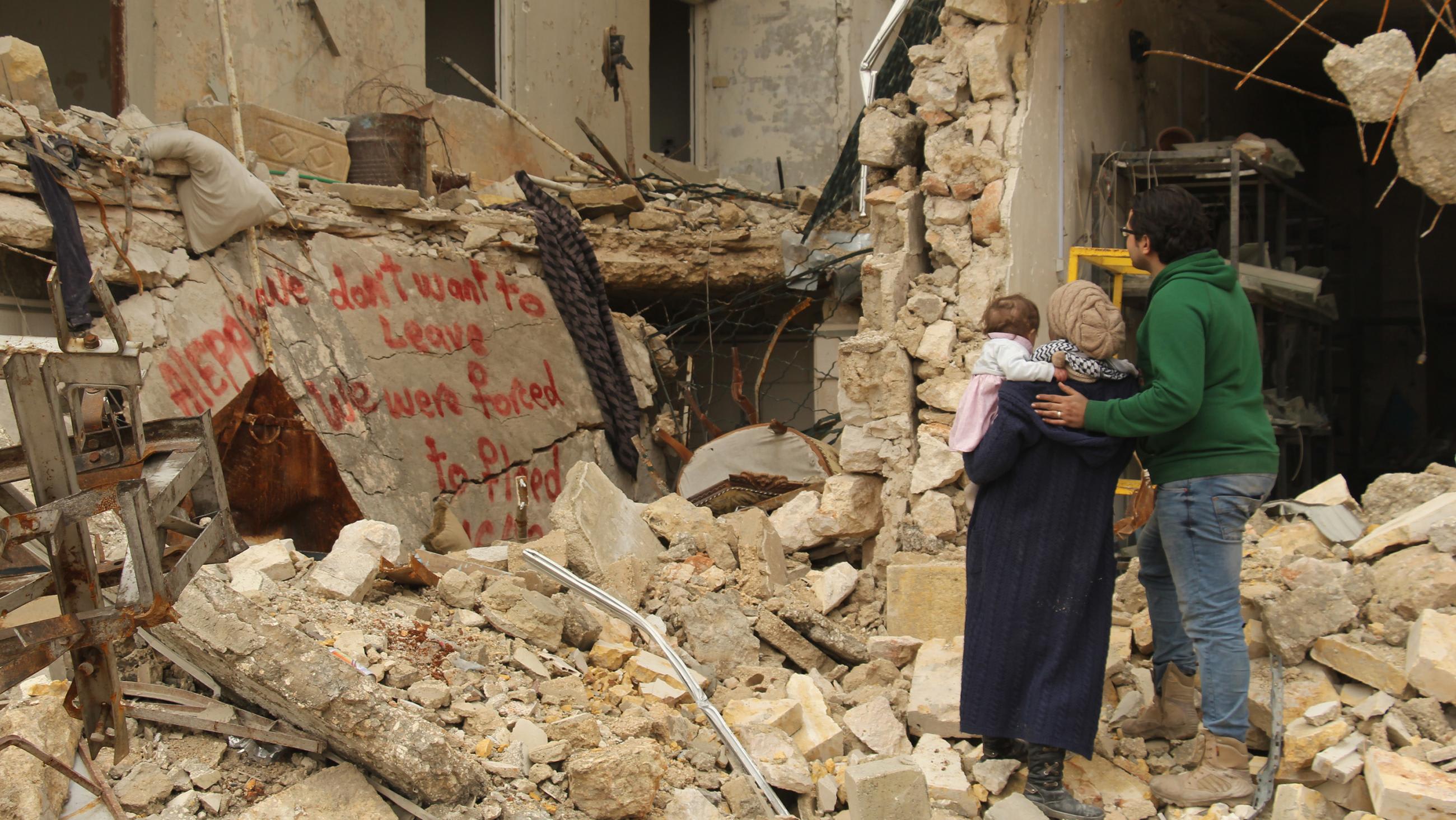 The picture shows the filmmaker holding her baby with her husband at her side. They are standing on top of rubble in front of a bombed-out building with twisted metal exposed and walls crumbled. Spray paint on a wall nearby reads,