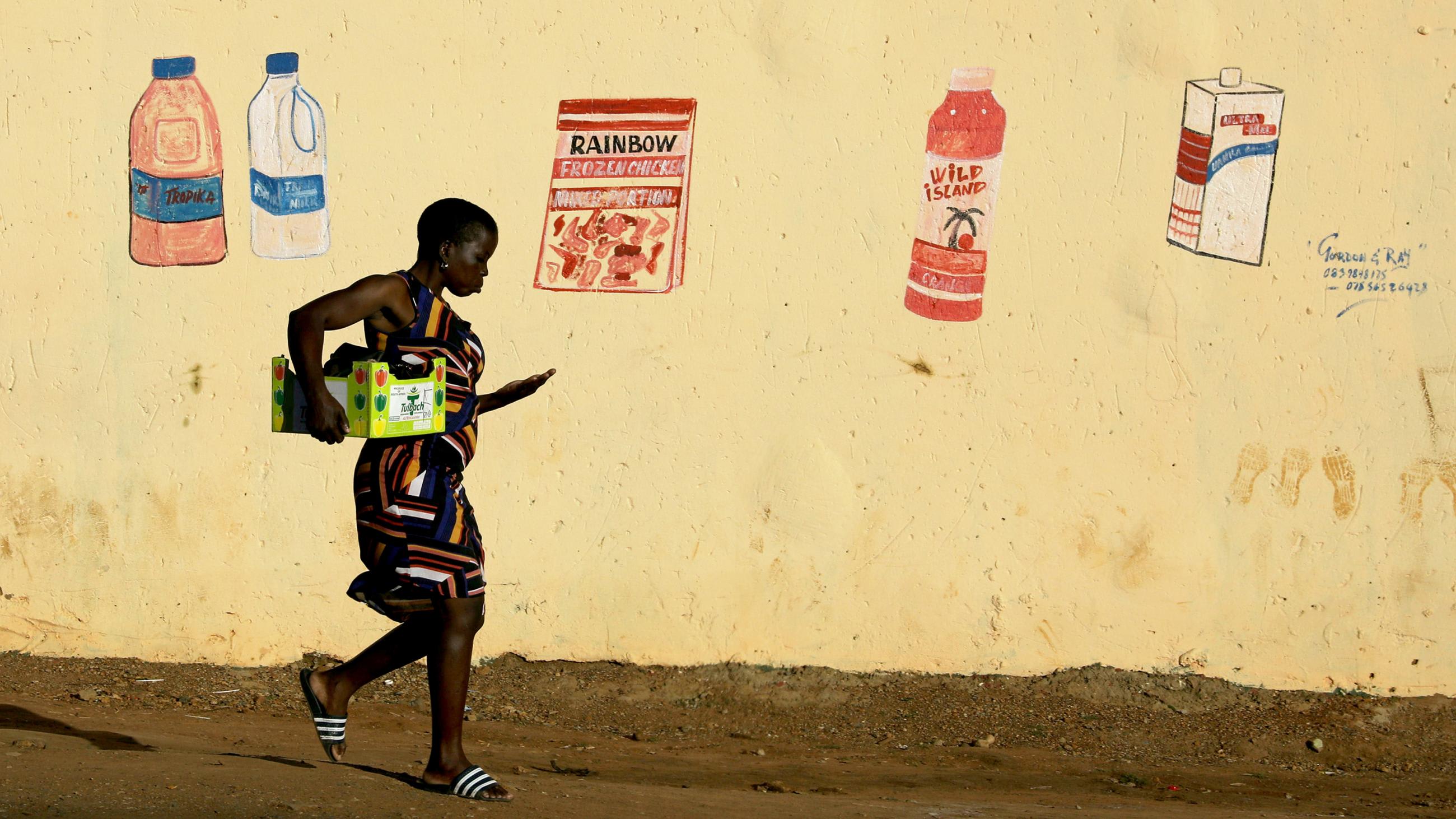 This is a striking photo of a woman carrying a box tucked under one arm and counting coins in the other walking in front of a butter yellow wall with faded, hand-drawn grocery items.