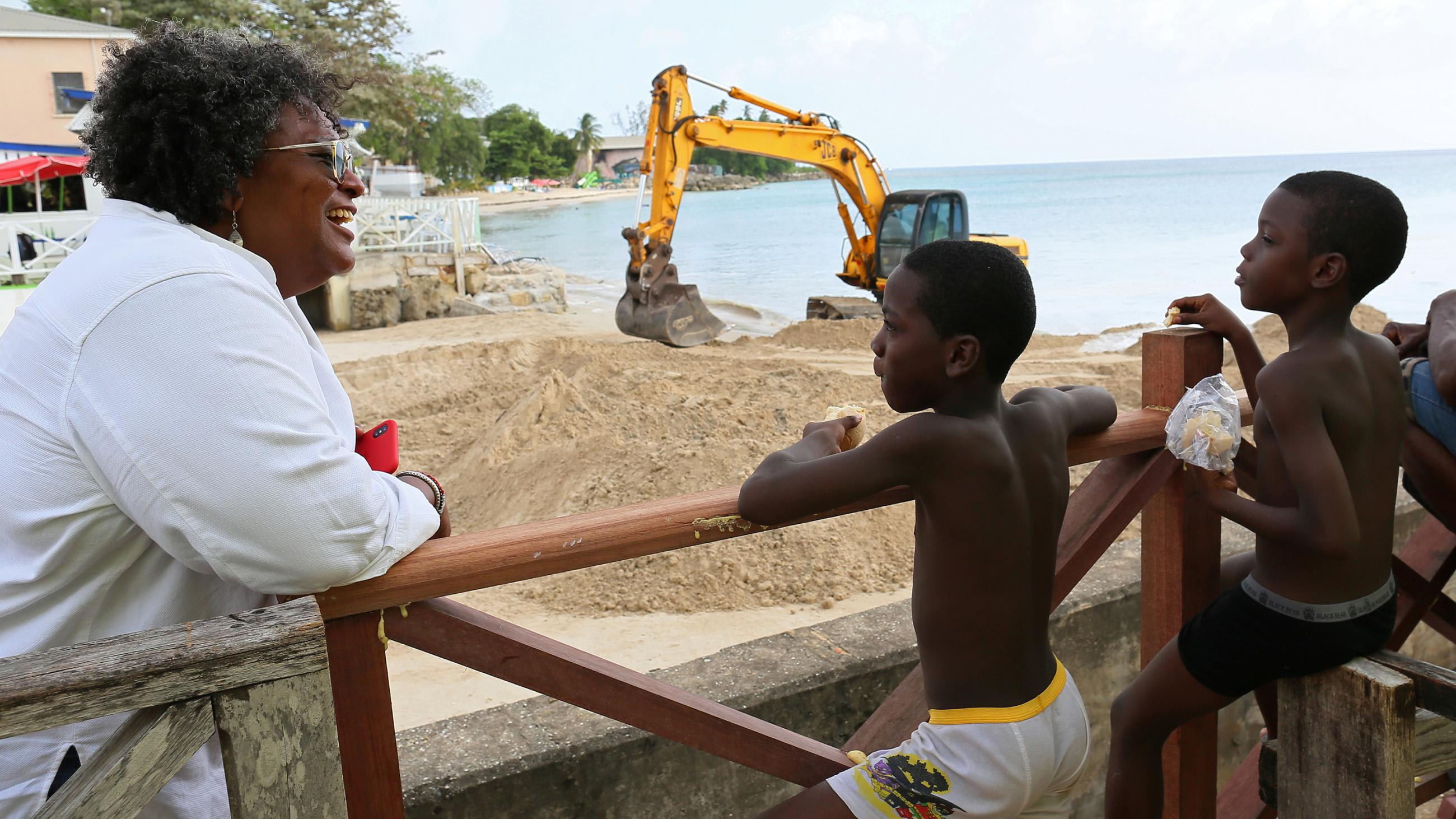 The photo shows the prime minister at the beach leaning on a rail talking to several young children. Construction machinery can be seen in the background.