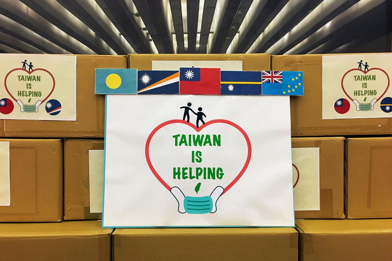 The photo shows boxes stacked and decorated with country flags and a large sign that reads, "Taiwan Is Helping."