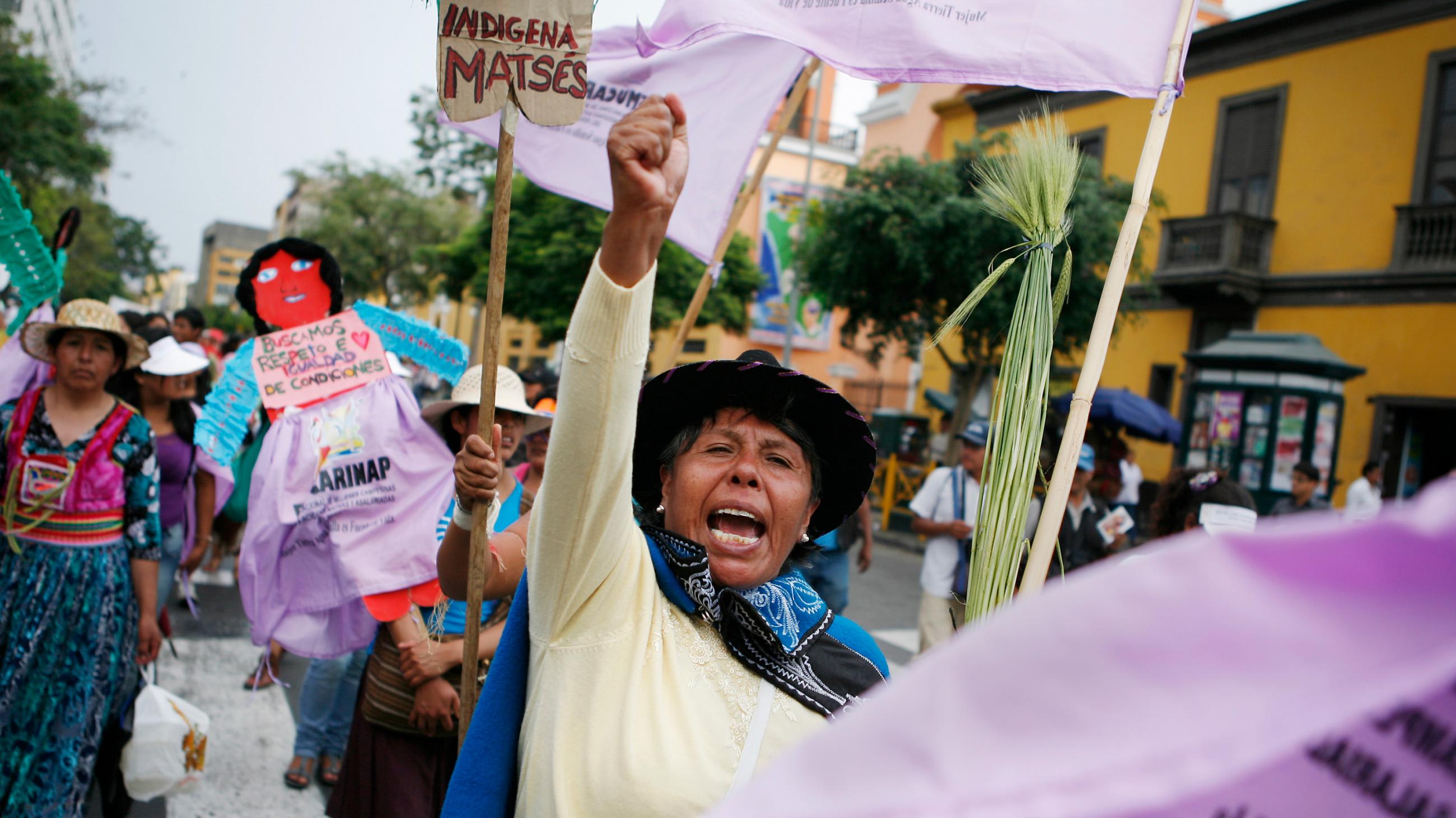 The photo shows a line of protesters marching down a street with a woman in the middle of the frame raising her hist and shouting.