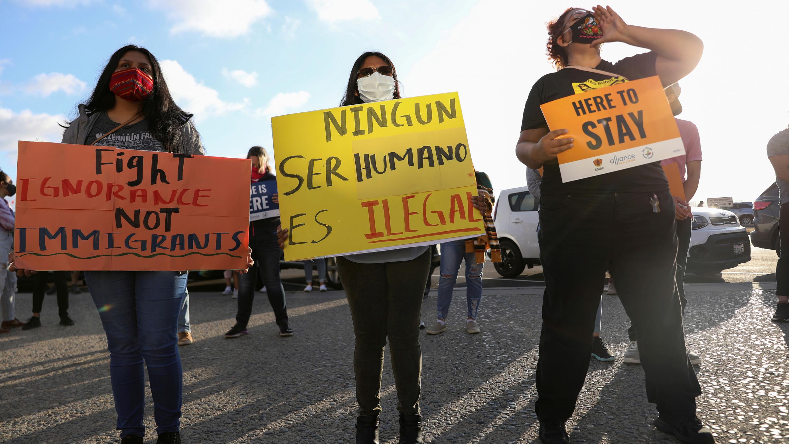 The photo shows three people holding handmade signs in a protest. The sign in the middle reads: "No human being is illegal."