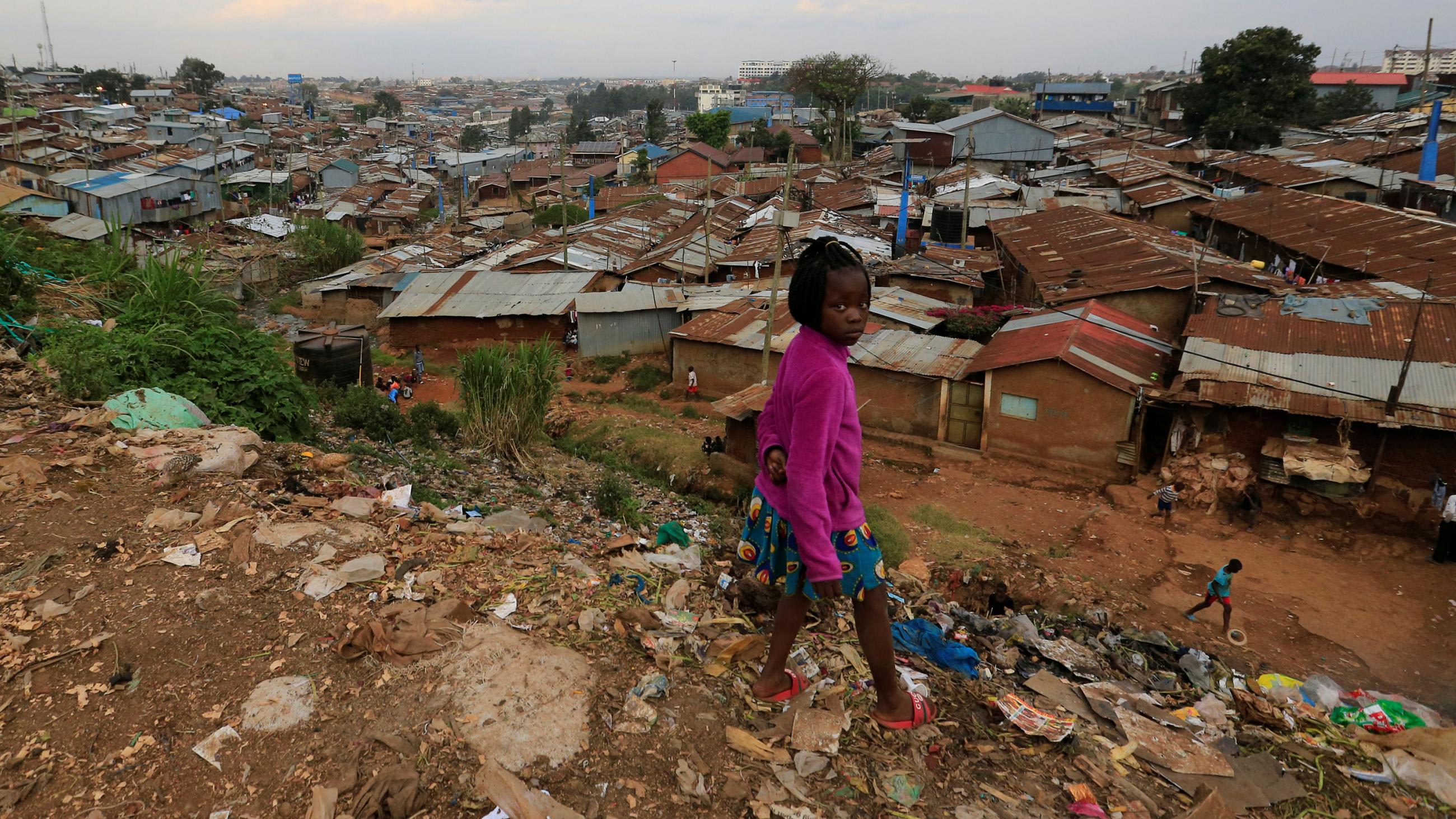 The photo shows a girl standing atop a hill littered with trash overlooking a residential area of poor houses with rusty metal roofing.