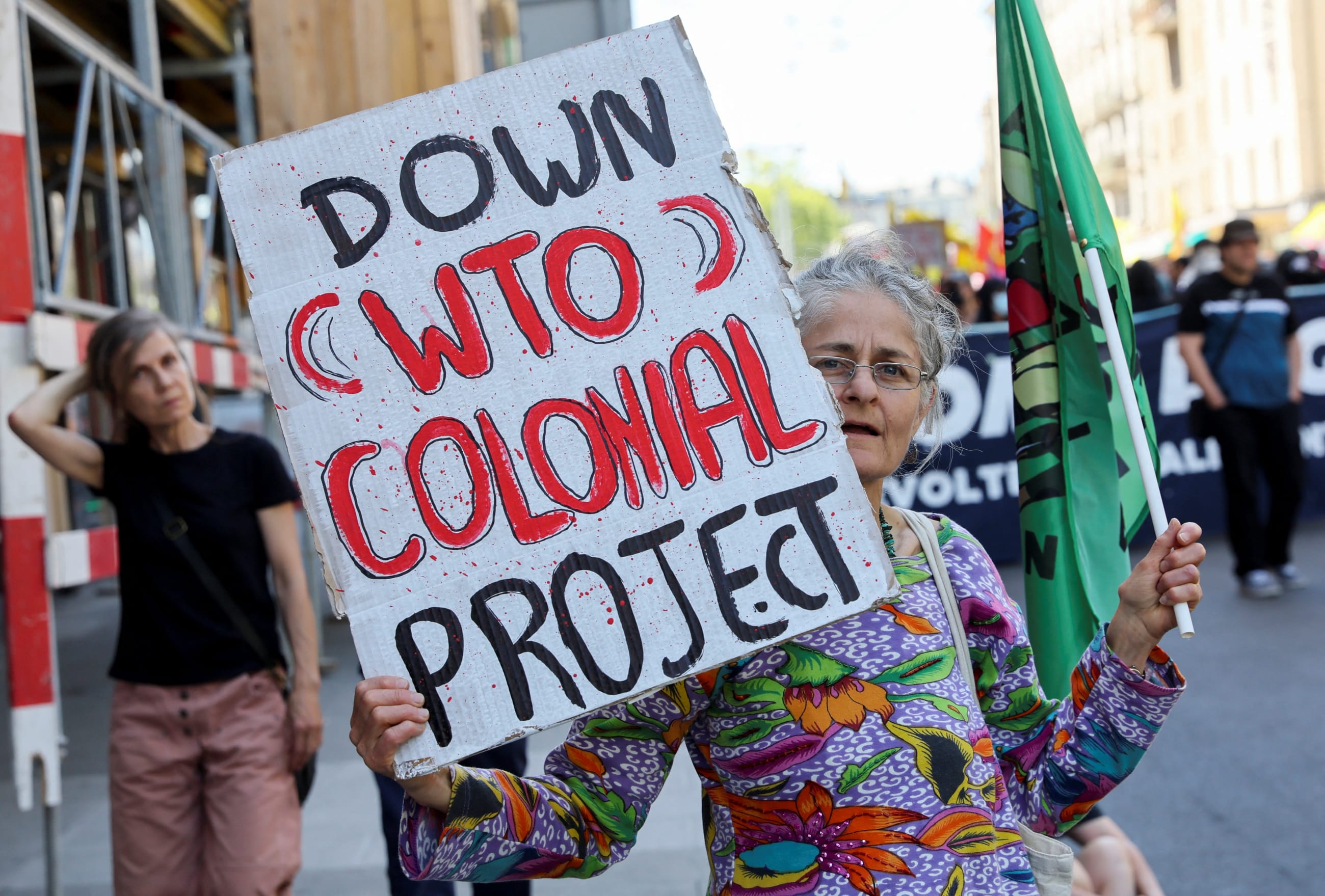 An older woman with white hair holds a  white-and-red sign that reads "Down WTO colonial project" in protest of the WTO ministerial conference in Geneva, Switzerland, on June 11, 2022.