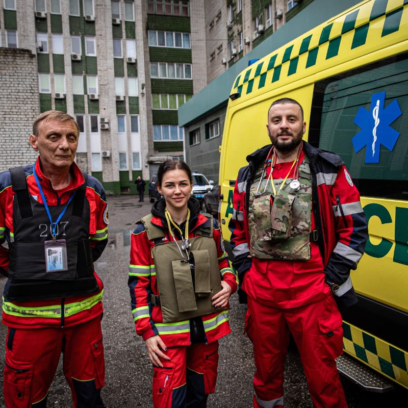 Health workers, two male and one female, wear red emergency outfits, stethoscopes, and bullet-proof vests as they  pose beside their yellow ambulance at a hospital in Kharkiv. 