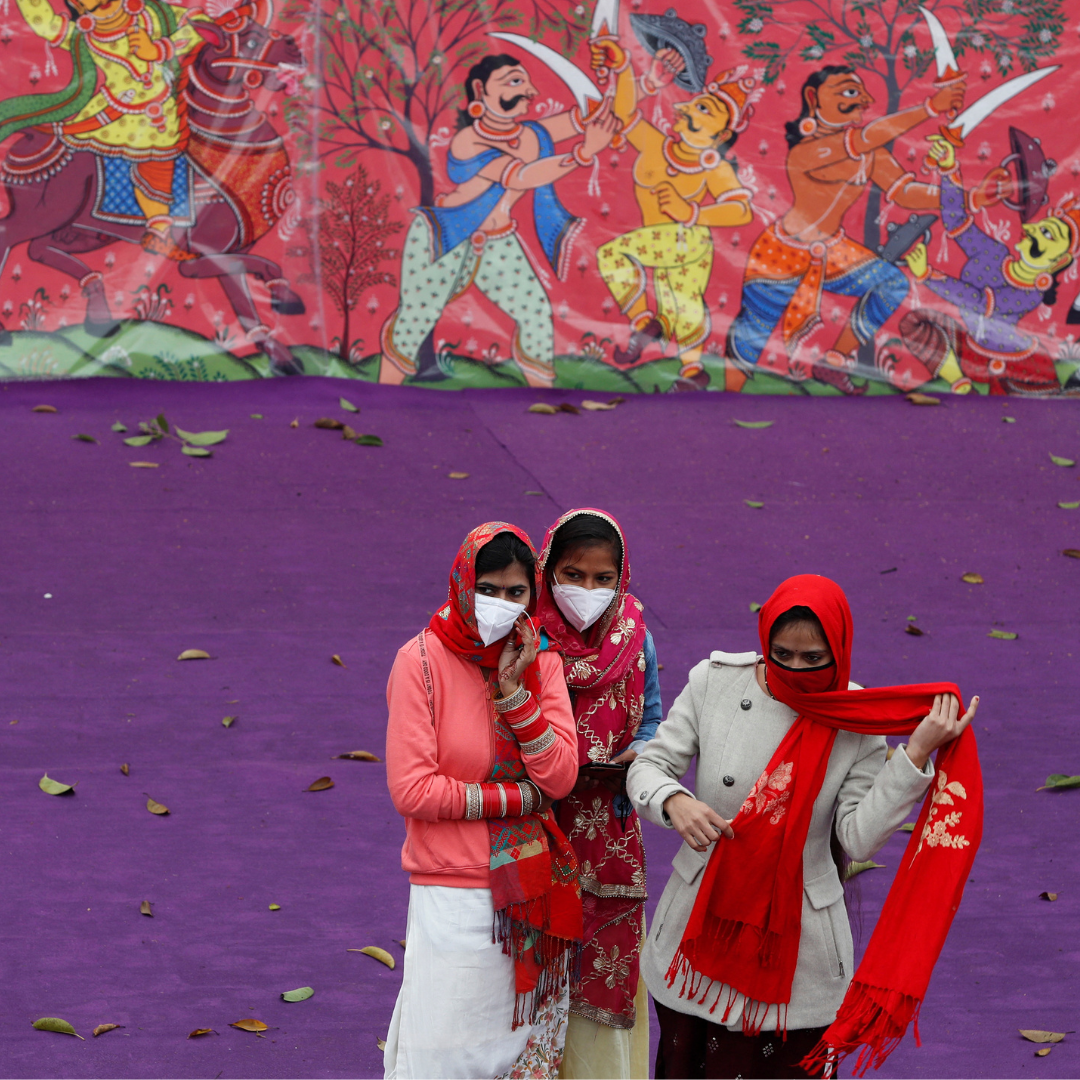 Women wearing protective face masks and colorful red and white saris stand in front of a purple wall and a color mural as they watch a dress rehearsal for the Republic Day parade, in New Delhi, India, on January 23, 2022. REUTERS/Adnan Abidi