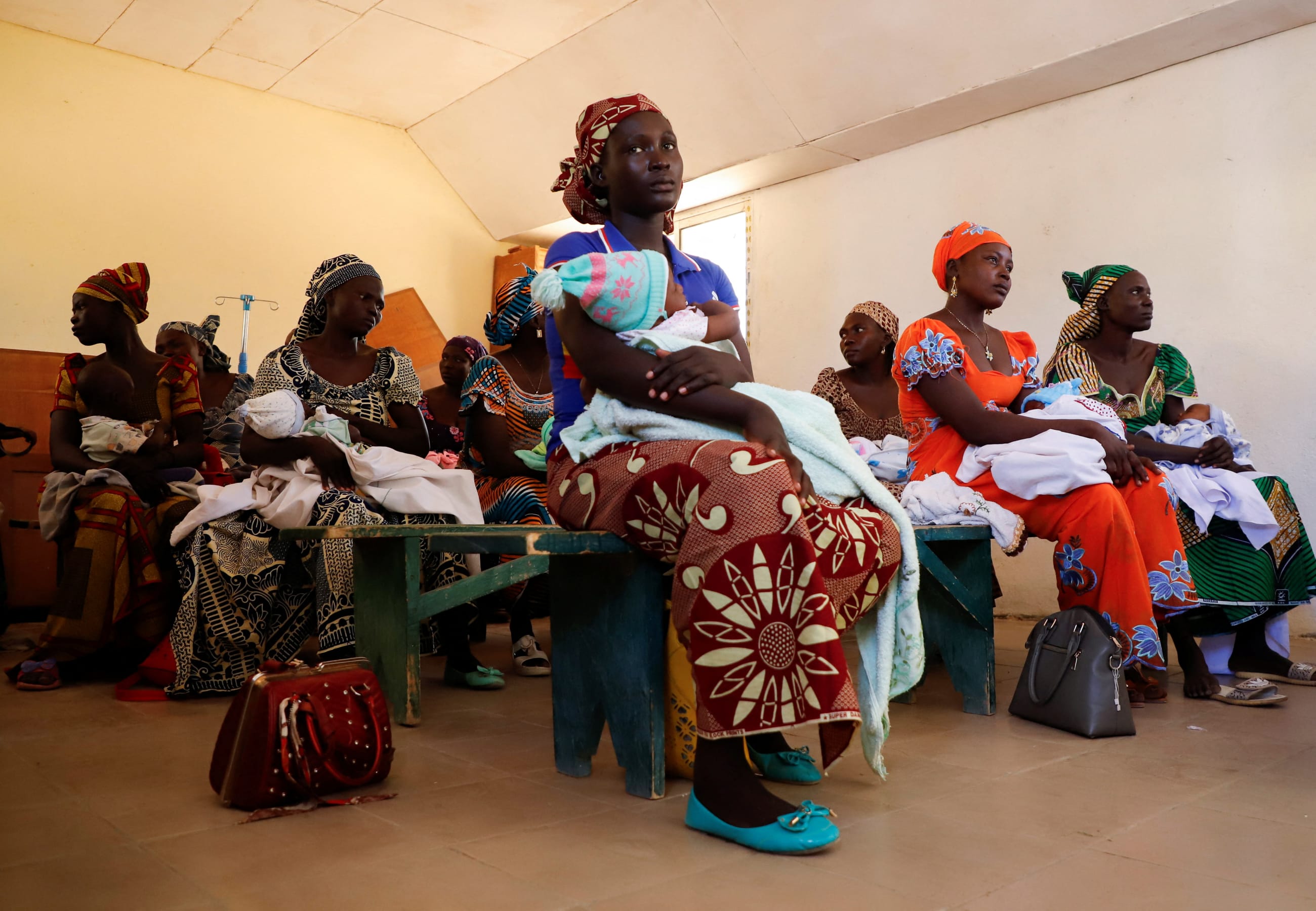 A group of women sit on benches inside a clinic, each holding an infant wrapped in blankets. They wear colorful dresses and headscarves, and handbags rest on the floor nearby. The women are mothers holding their children as they wait to be vaccinated against malaria, at a health center in Datcheka, Cameroon, on January 22, 2024.