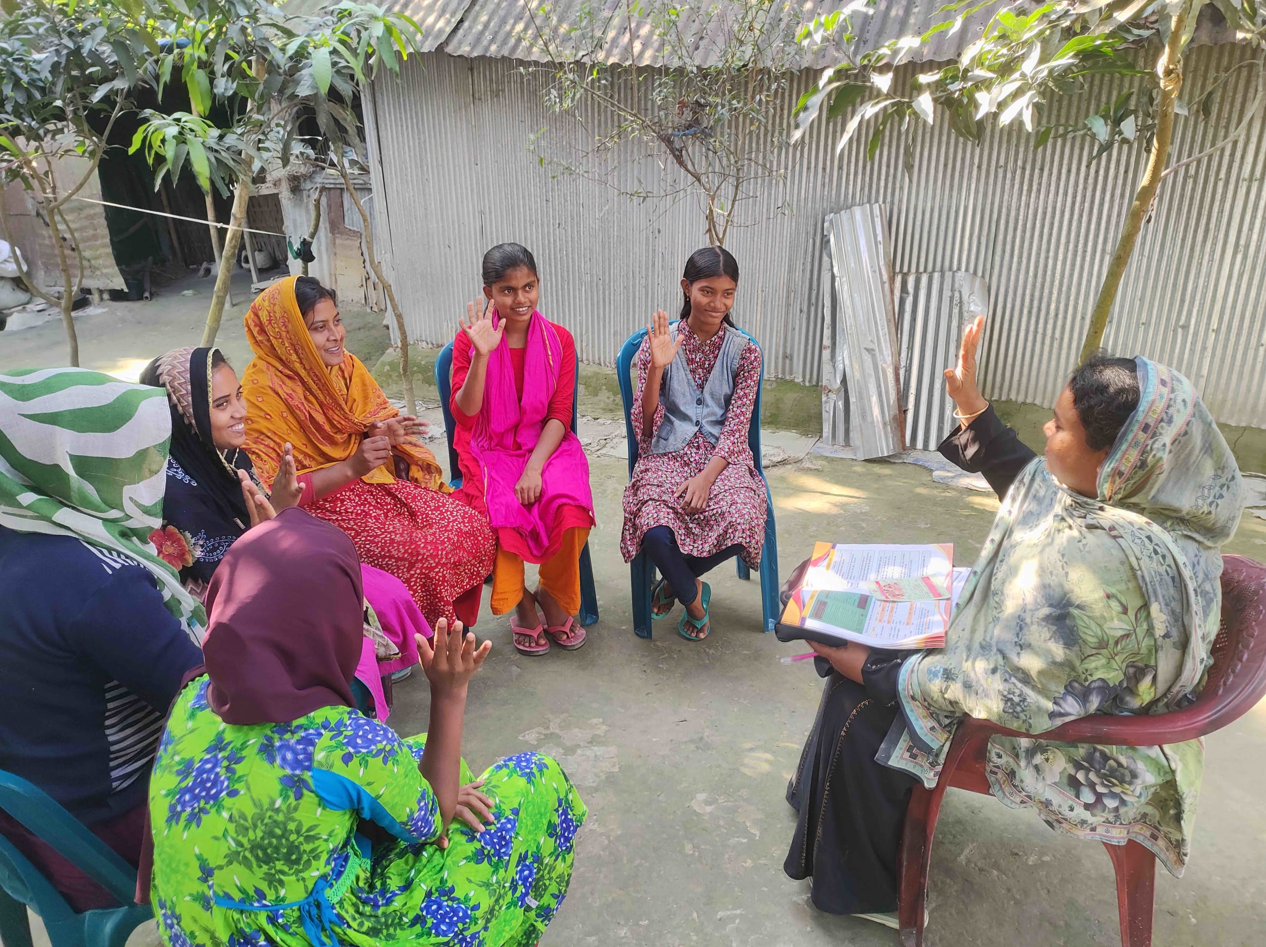 A group of adolescent girls and women sit in a circle outdoors as a health leader in a patterned scarf leads a training session, holding up her hand while participants raise theirs in response.