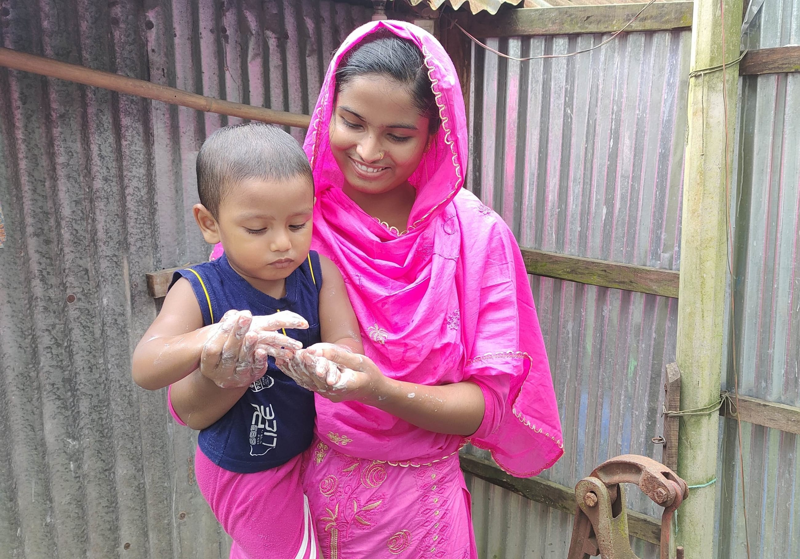 Health leader Sabikunnahar, wearing a bright pink traditional outfit, smiles while helping a young child wash their hands with soap and water outdoors beside a corrugated tin wall.
