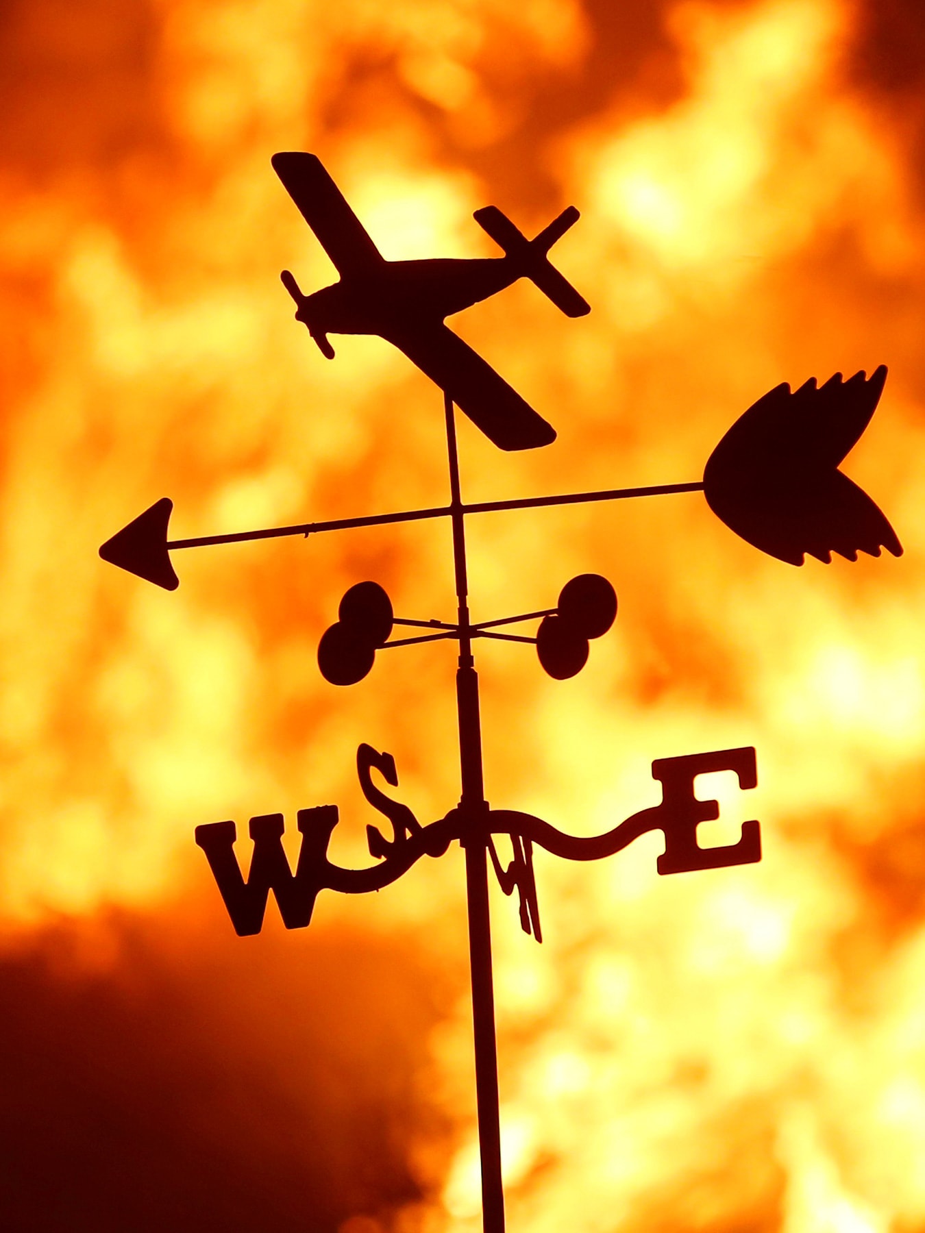 Picture shows a thin metal toy-like vain with a plane, and arrow and a “NESW” directional indicator silhouetted against a burning fire.