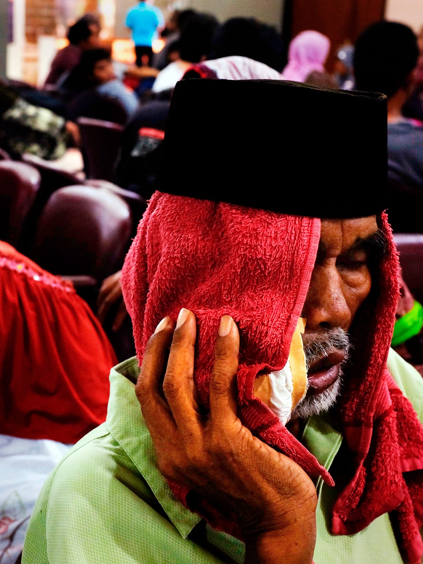 The picture shows a crowded waiting room with a man in the foreground with a red cloth draped over his head and holding what appears to be a bag of ice against his cheek. He looks like he is suffering.