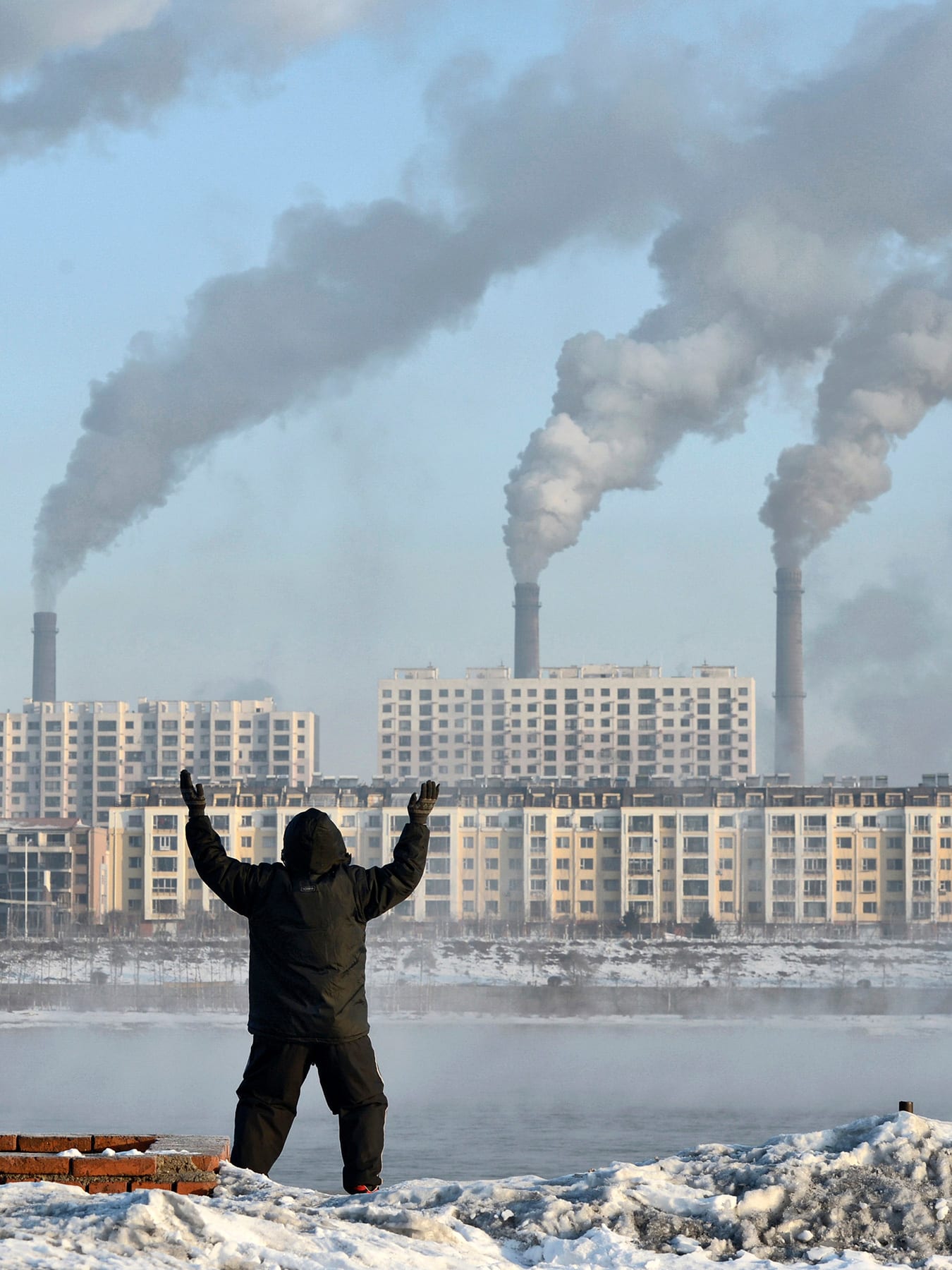 The photo shows the man in the foreground, back to the camera, with buildings on the other side of the river pouring out smoke.
