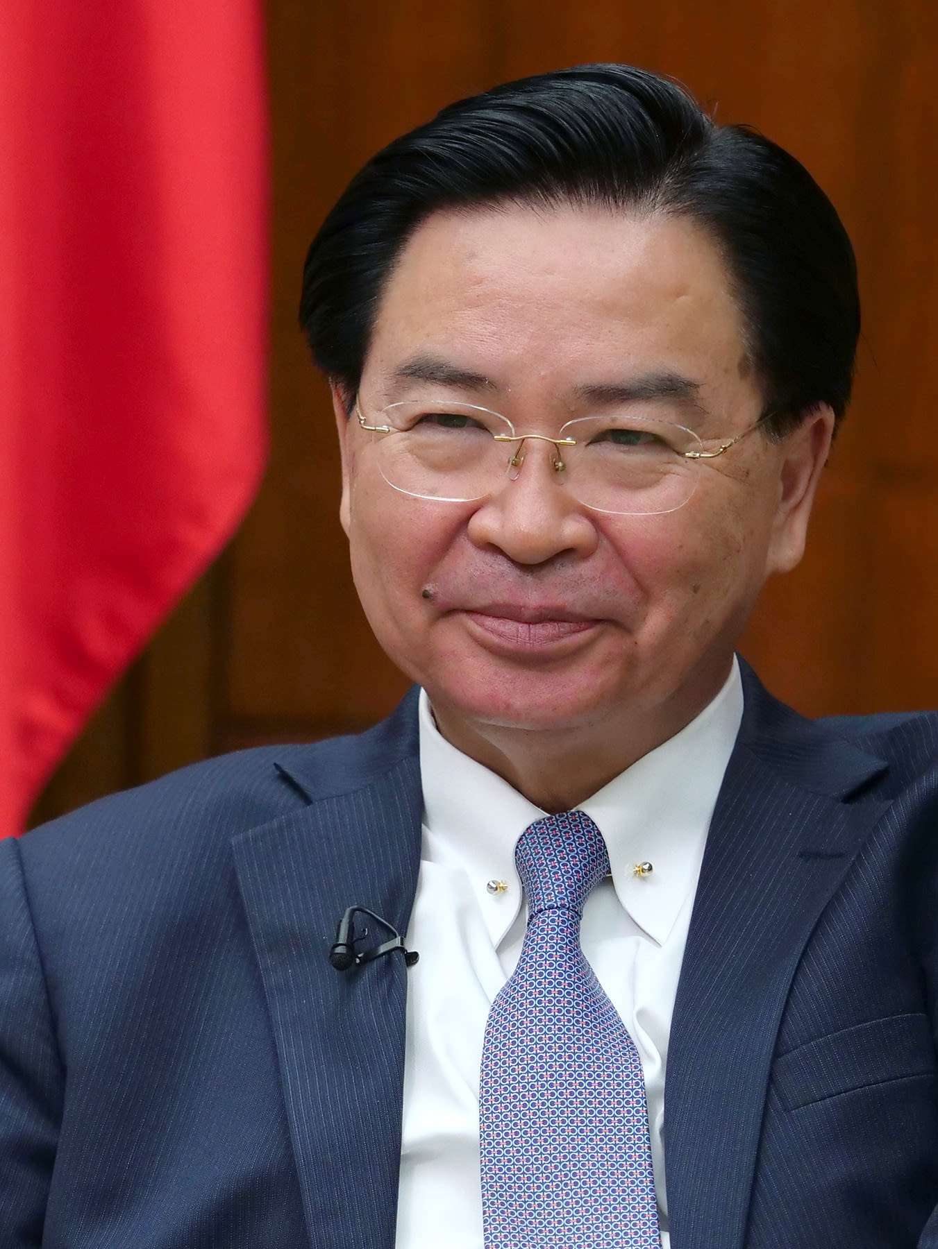 The photo shows the minister seated in a comfortable chair looking off-camera and smiling.