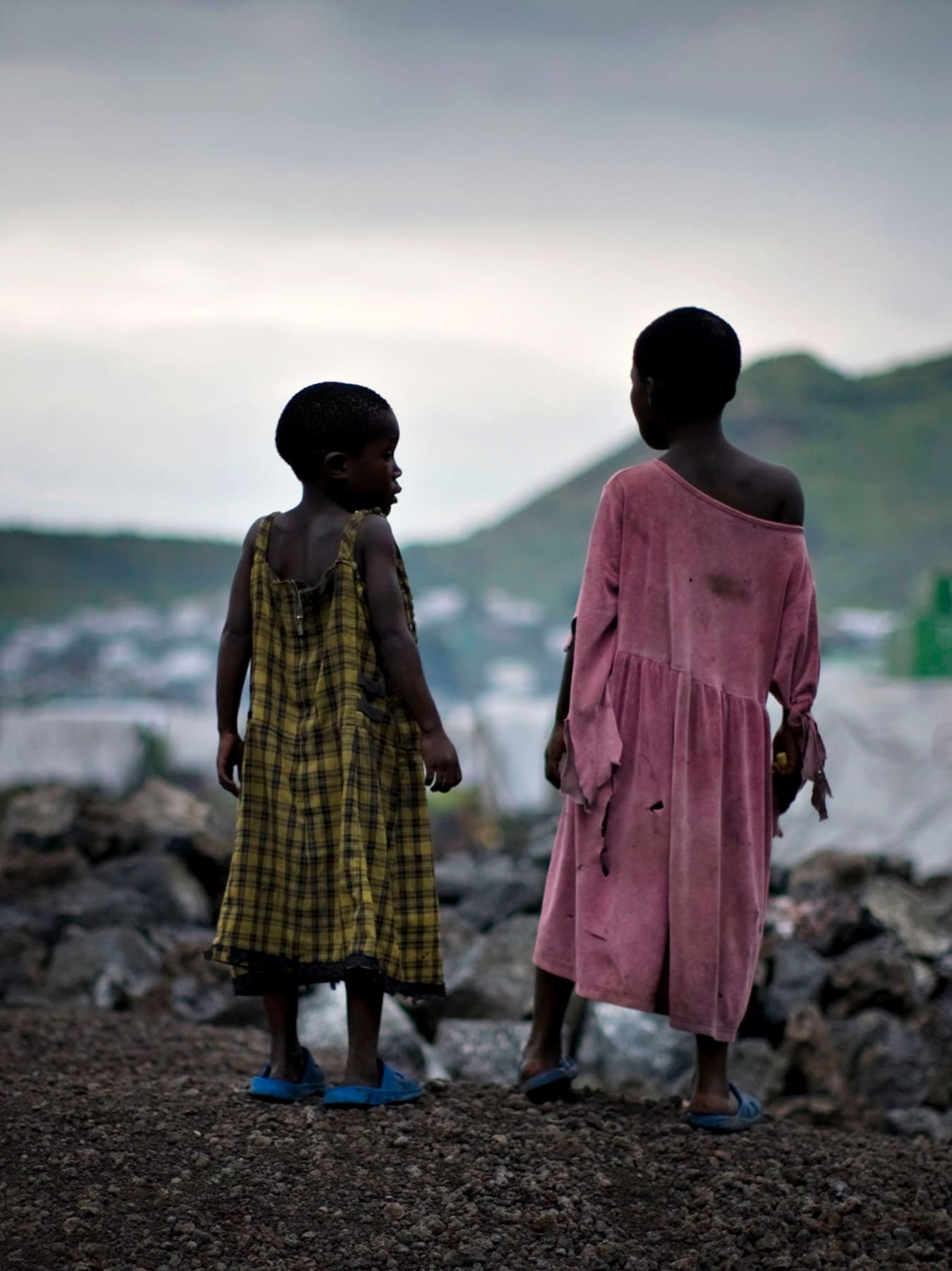 The photo shows two girls standing on a hill overlooking a camp filled with white tents at sunset.