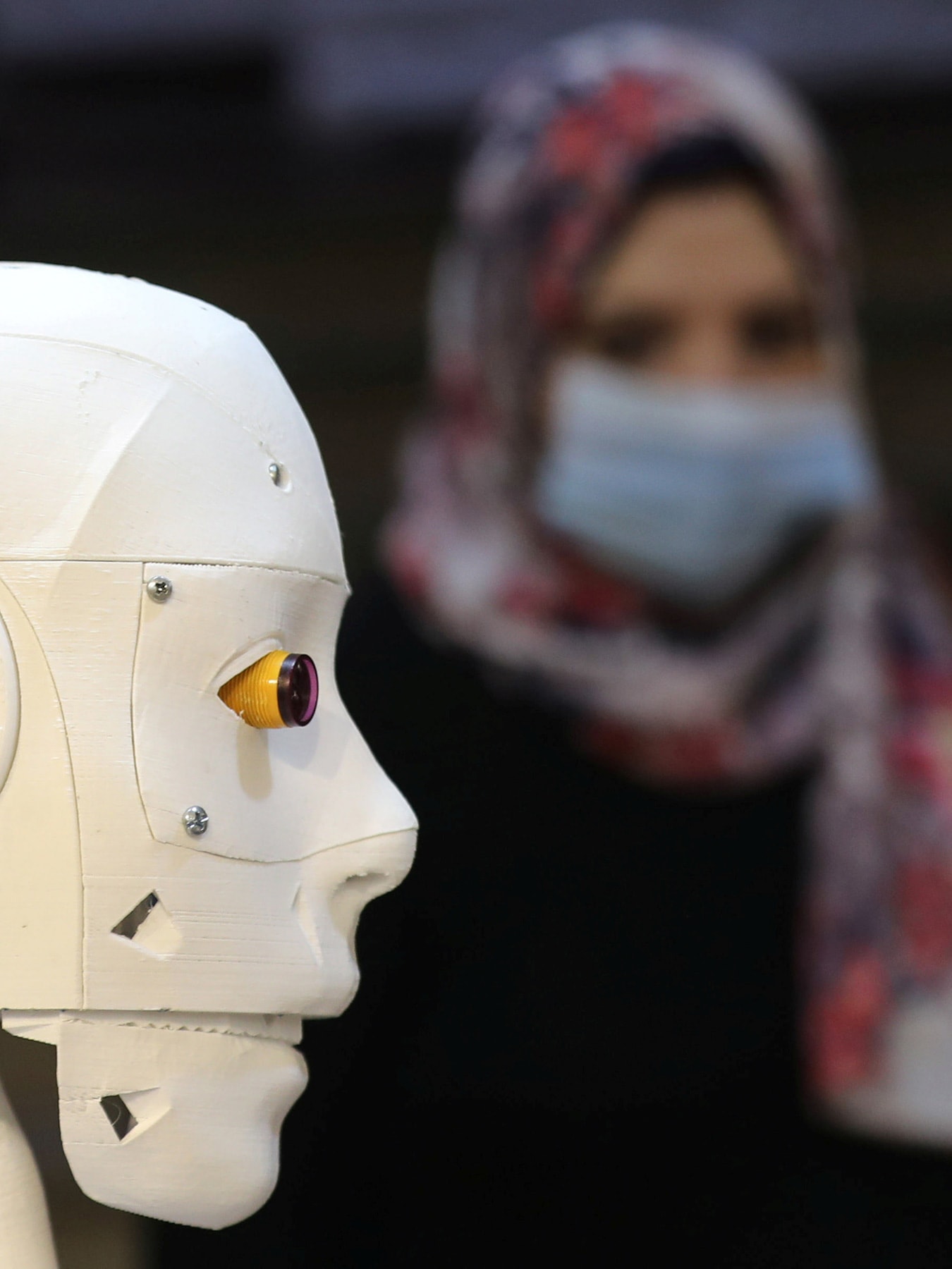Picture shows the robot in the foreground while a woman in the background wearing a mask looks on.