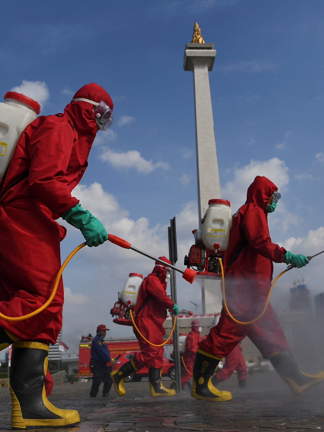 Photo shows several workers in bright red protective suits walking though an open area spraying from tanks they carry on their backs. A monument can be seen in the background.