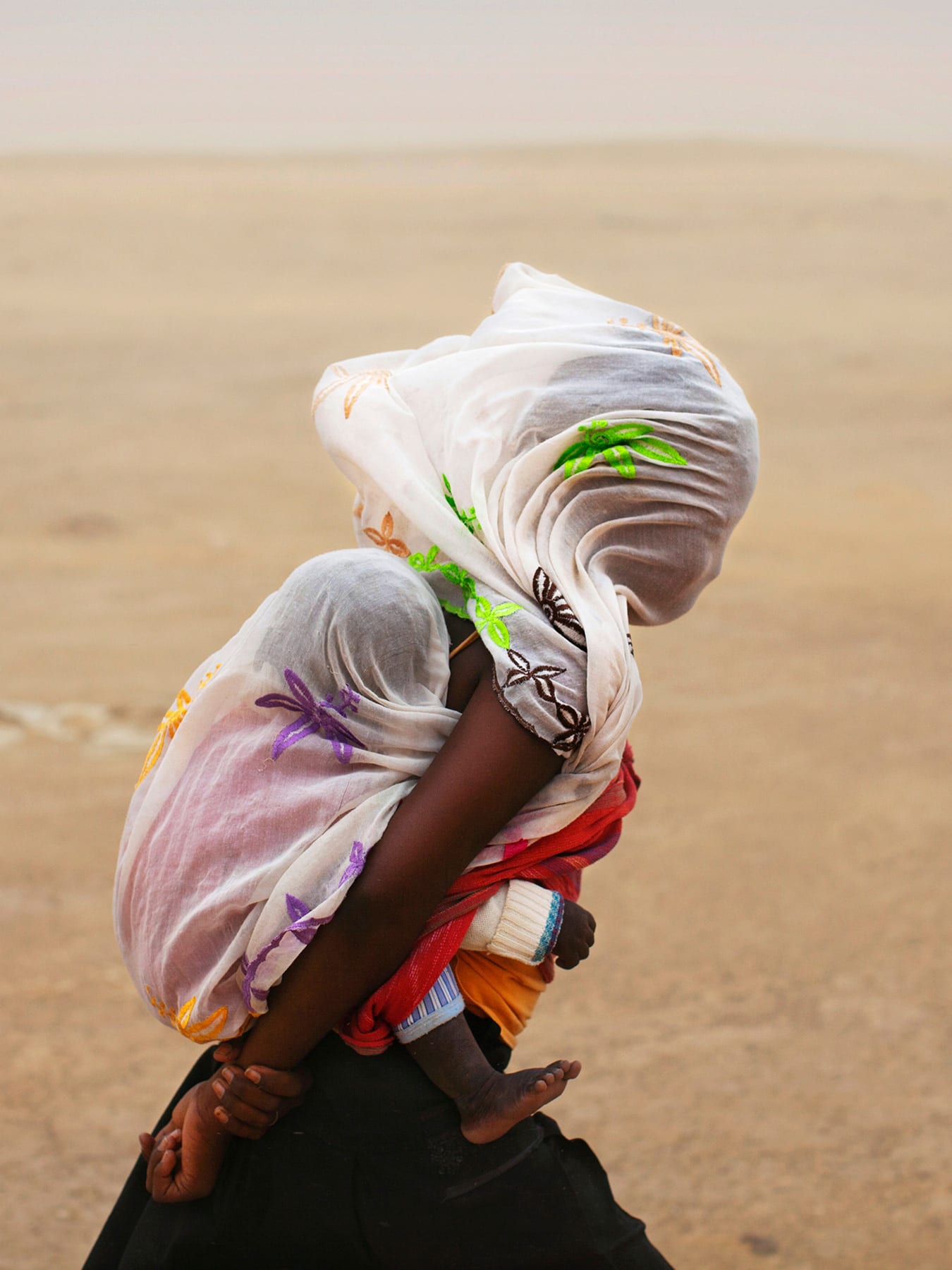 The photo shows a woman with a baby on her back walking through a desert. The wind is blowing strongly, as evidenced by the billowing cloth on their heads.