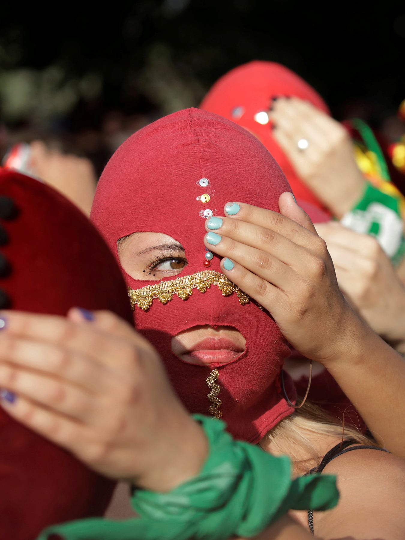 The photo is a striking image of women wearing red masks lined up, each person holding a hand over one eye of the person in front of them. 