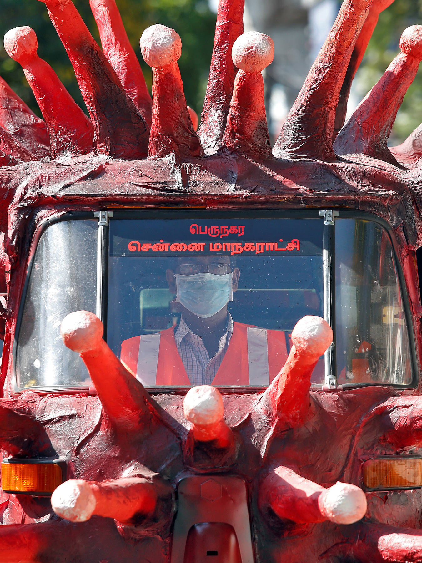 The image shows a vehicle painted red and covered with crown spikes reminiscent of the virus, which appear to be made of paper mache.