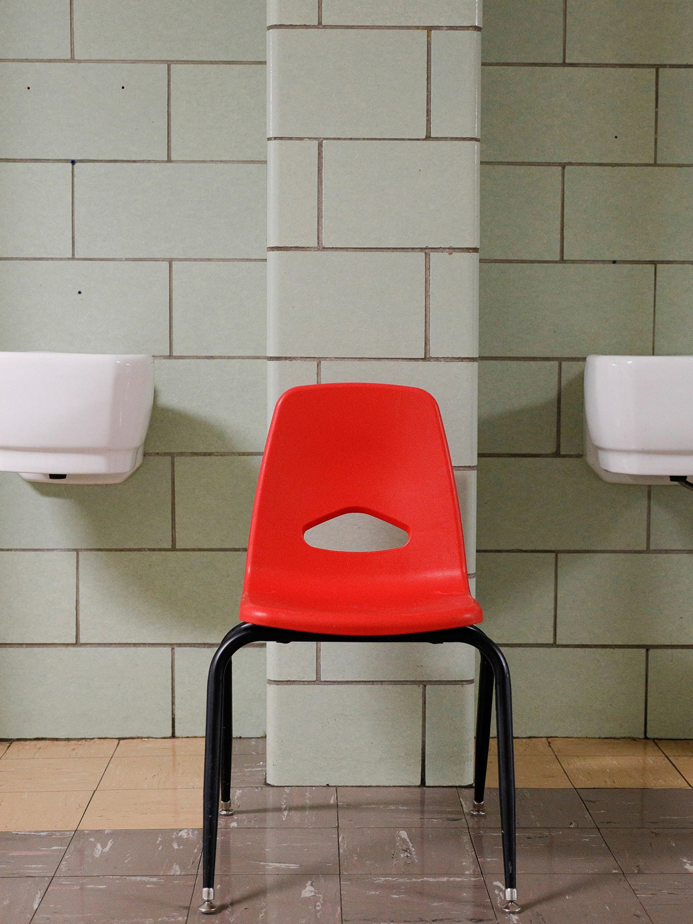The photo shows a red chair in a beige-colorored hallway where two porticos feature white porcelain basins now dry for the pandemic. 