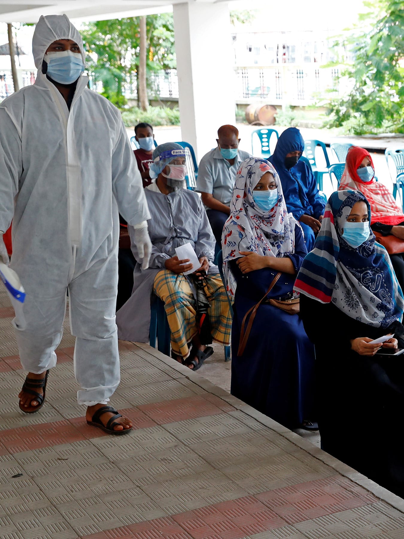 The photo shows a crowd of people wearing masks waiting on line in chairs while a health worker in full protective gear walks beside them.