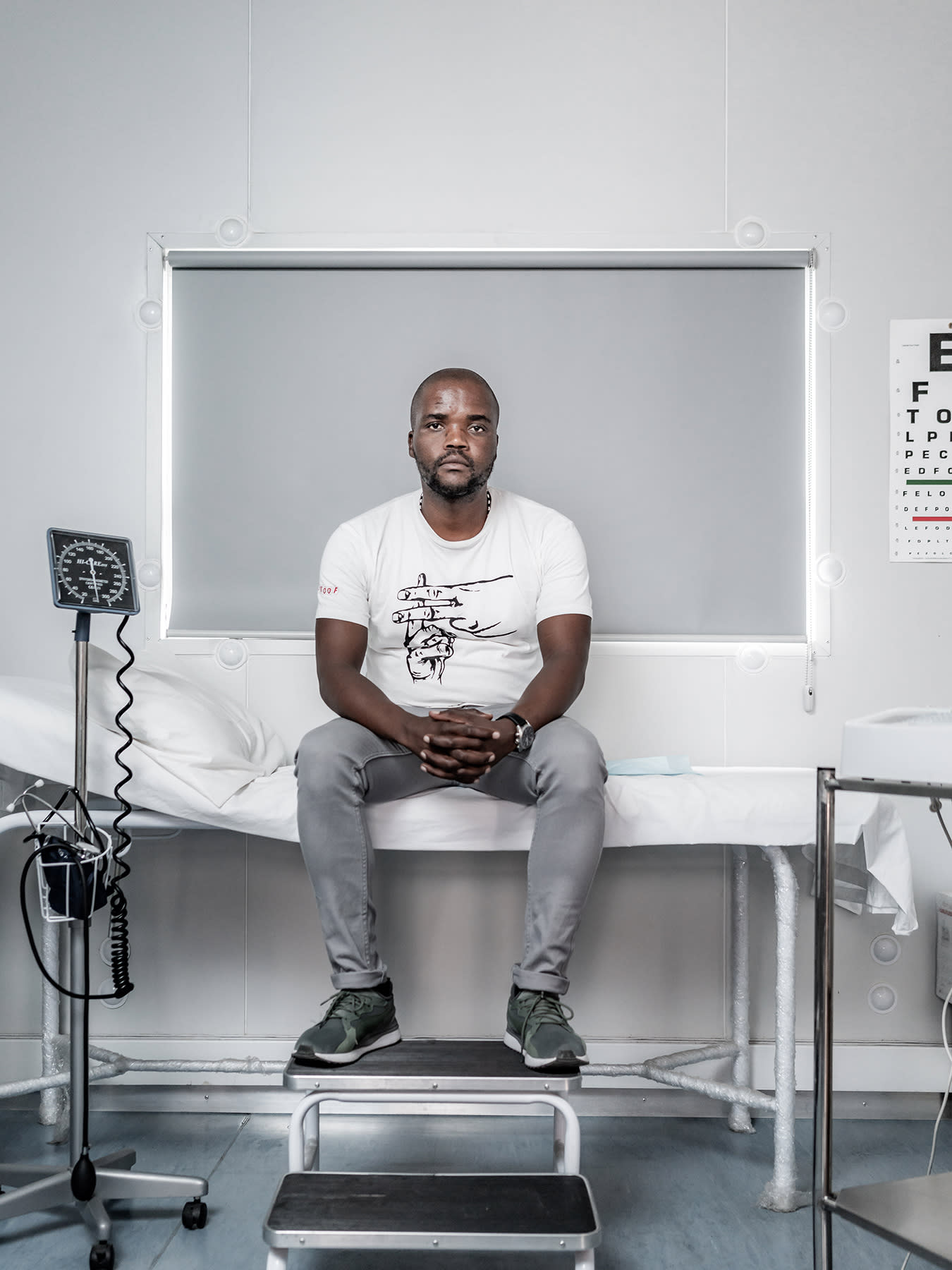 The photo shows a man sitting in a clinic with various monitors and equipment. An eye exam is visible on the wall. 