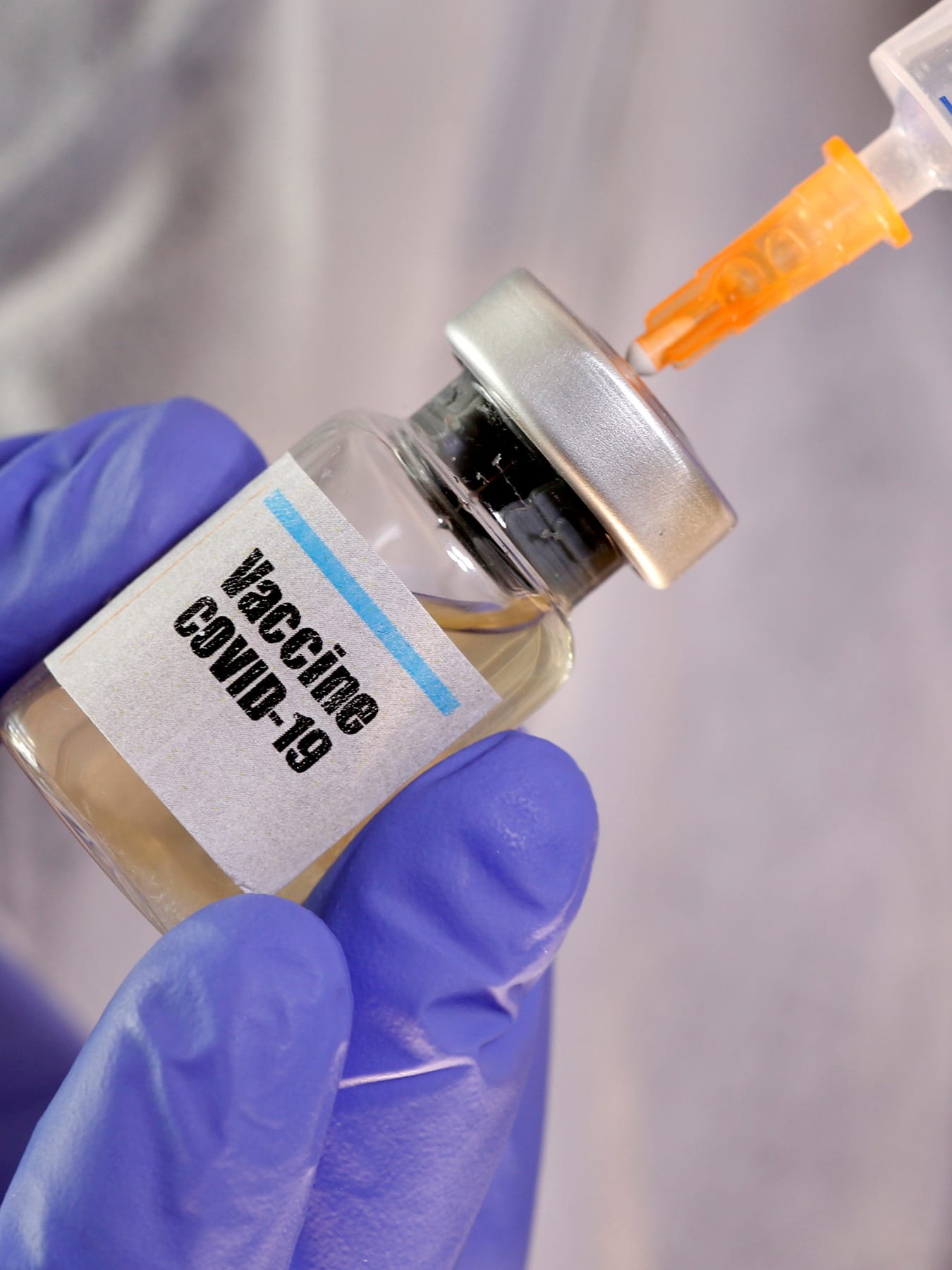 The photo shows a woman holding a small vial to her face. She is wearing a mask and gloves and the vial reads, COVID-19.