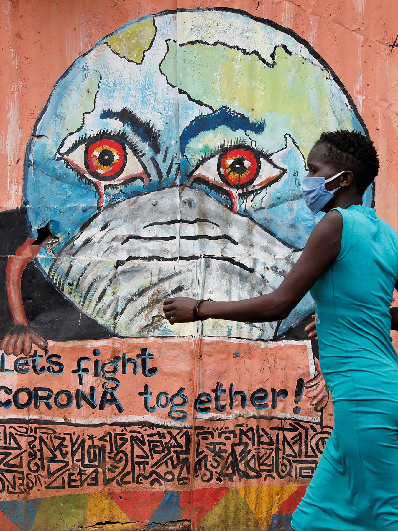 This is a striking photo of a woman in a blue dress wearing a facemask walking past a peach-colored wall with a graffiti of the Earth, also wearing a facemask, with bloodshot eyes and crying.