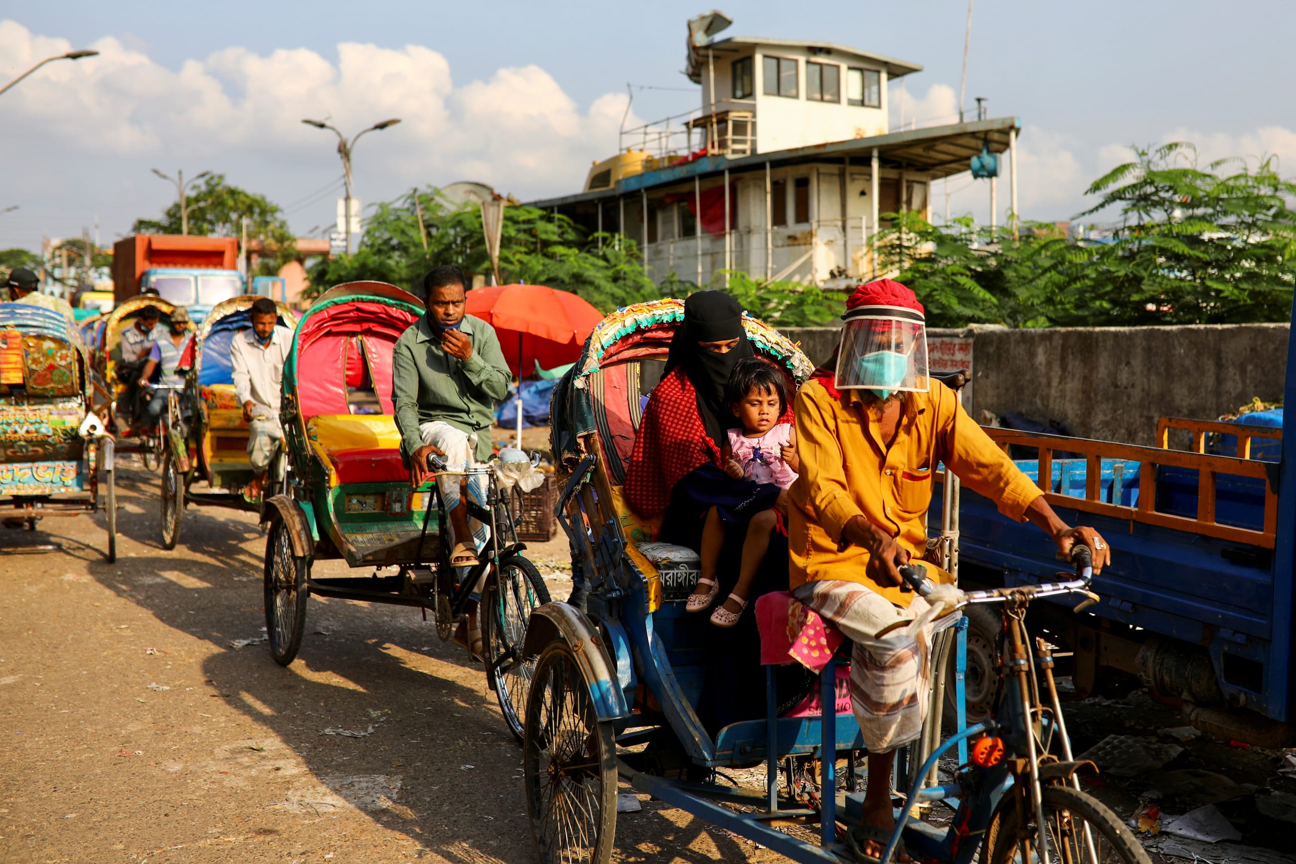 Ricksha pullers wearing face shields and blue medical masks carry passengers in a row rickshas with yellow, orange, and green covers amid the COVID-19 outbreak, in Dhaka, Bangladesh, on June 1, 2020.