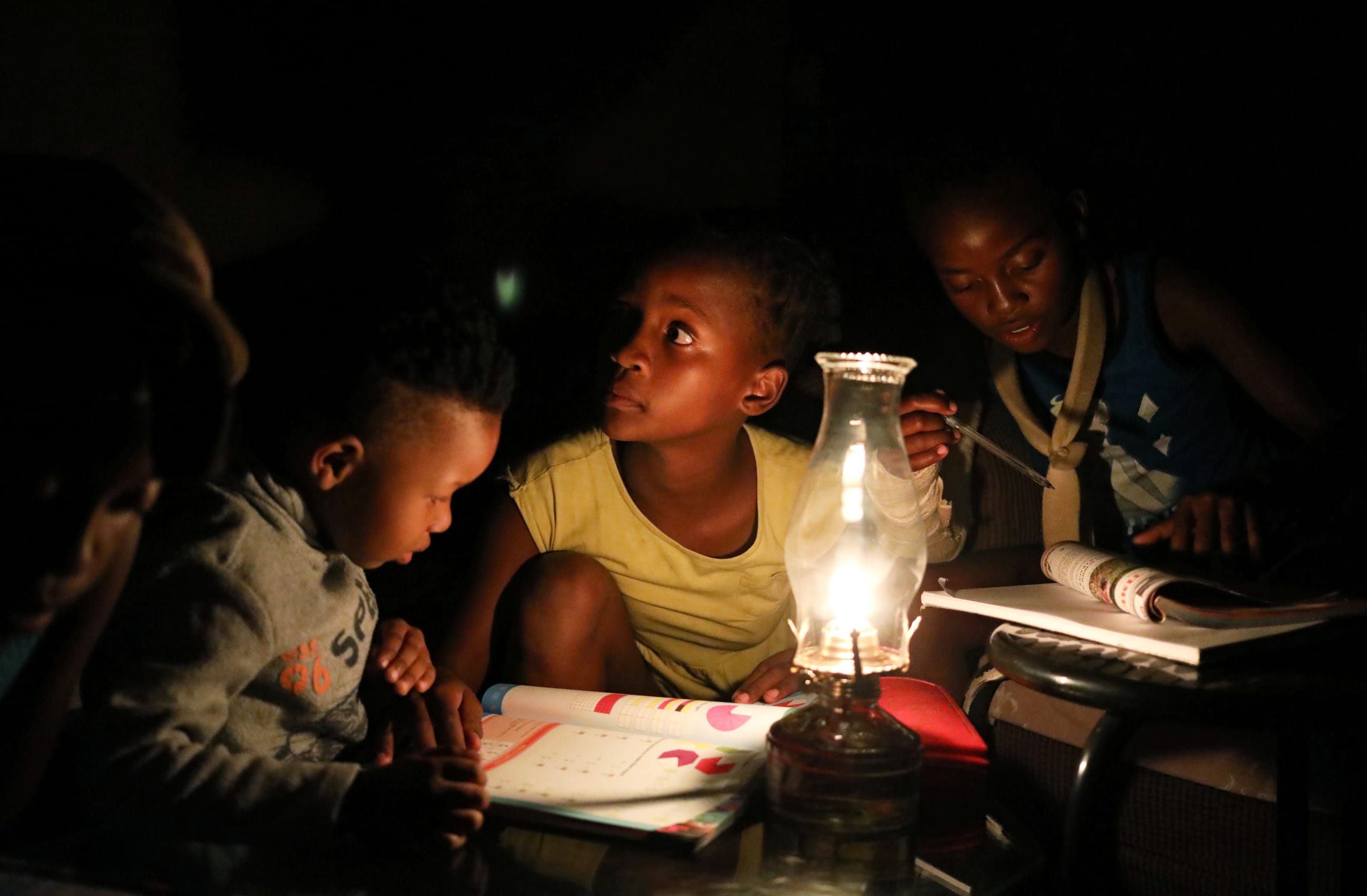 Three South African children sit in a dark room look on as they use a parafin light to study during an electricity load-shedding blackout in Soweto, South Africa, on March 18, 2021.