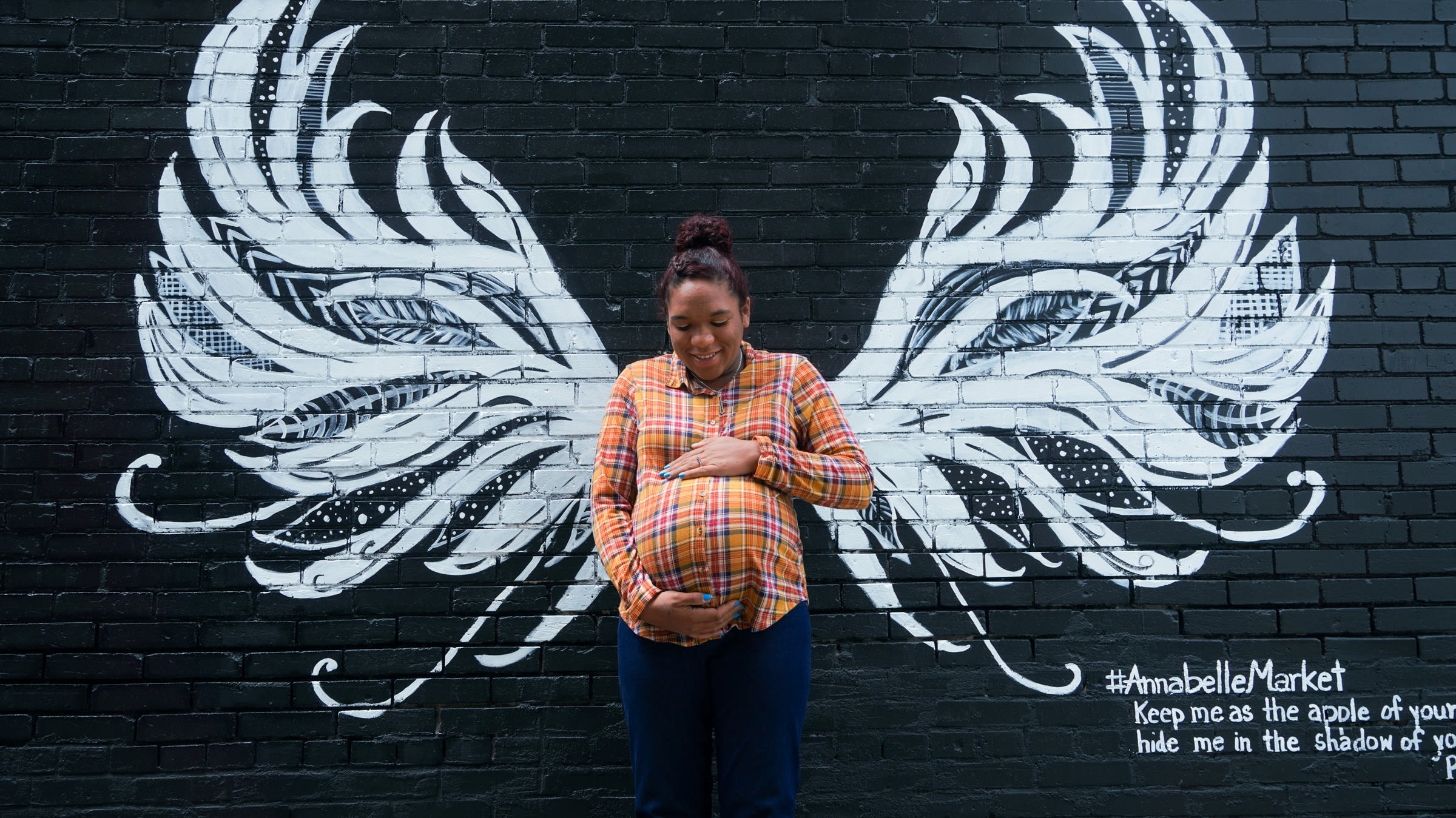 A Black mother in an orange shirt lovingly holds her pregnant belly in front of a mural on a black brick wall that gives her white angel wings in Anna, Illinois, U.S., May 10, 2021.