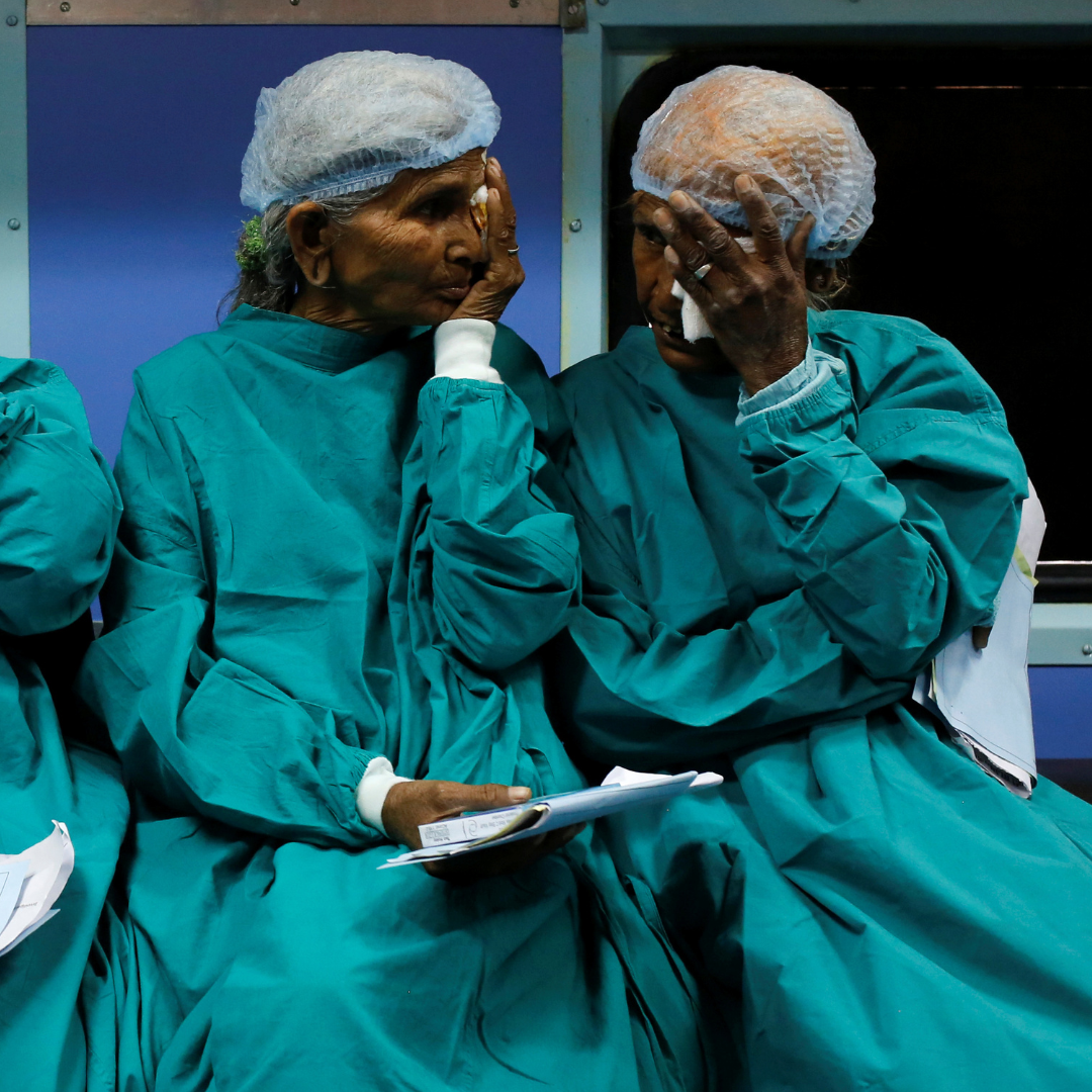 Two patients dressed in green medical gowns and plastics caps sit on a bench and cover their eyes as they wait for cataract surgery on the Lifeline Express, a hospital built inside a seven-coach train, at a railway station in Jalore, India, on March 31, 2018.