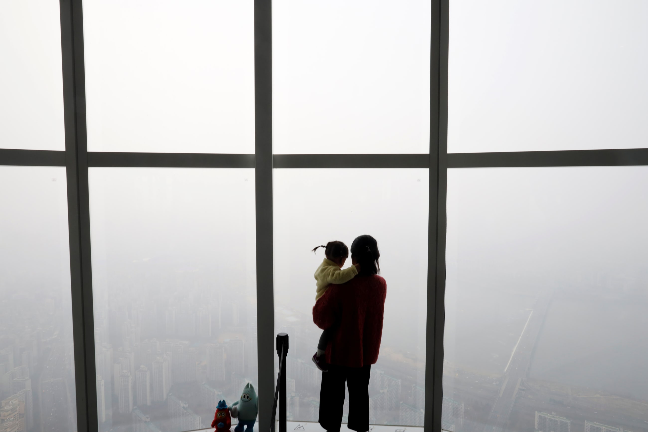 A woman holding her baby in her arms looks out a window at the city shrouded by a fine dust during a polluted day. 