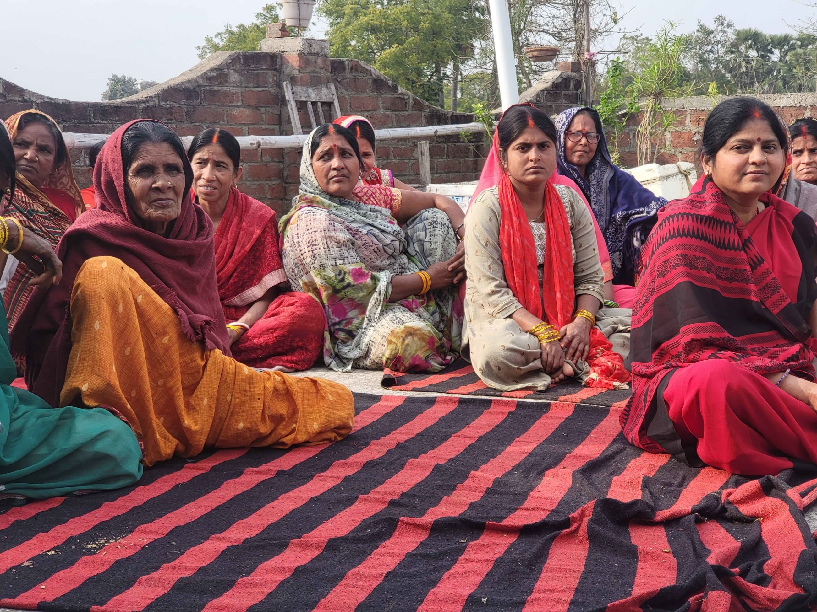 A group of women in colorful saris sit together on a striped mat outdoors, listening attentively during a community health education session.