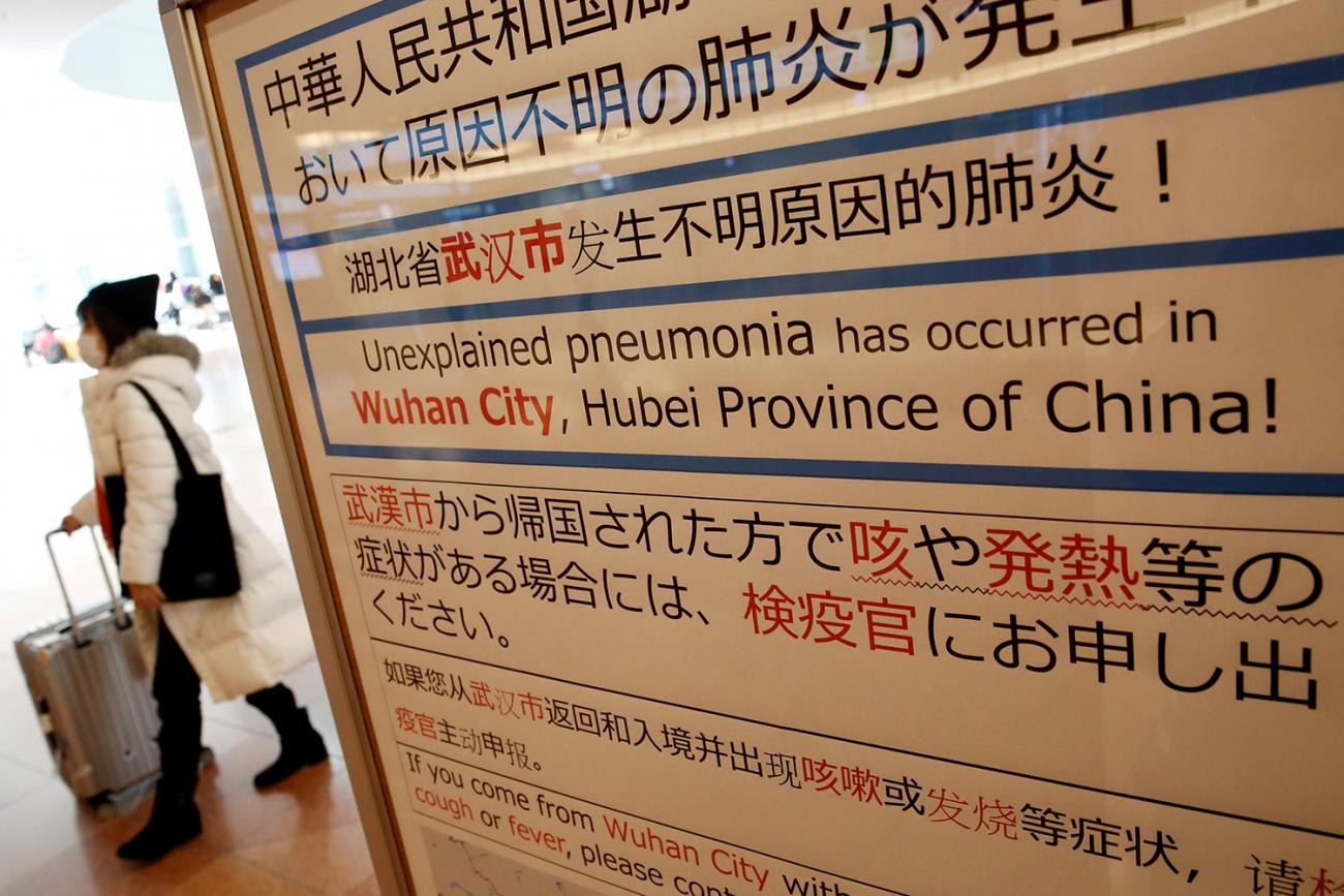 The photo shows a woman in a mask walking past a sign warning travelers in several languages.