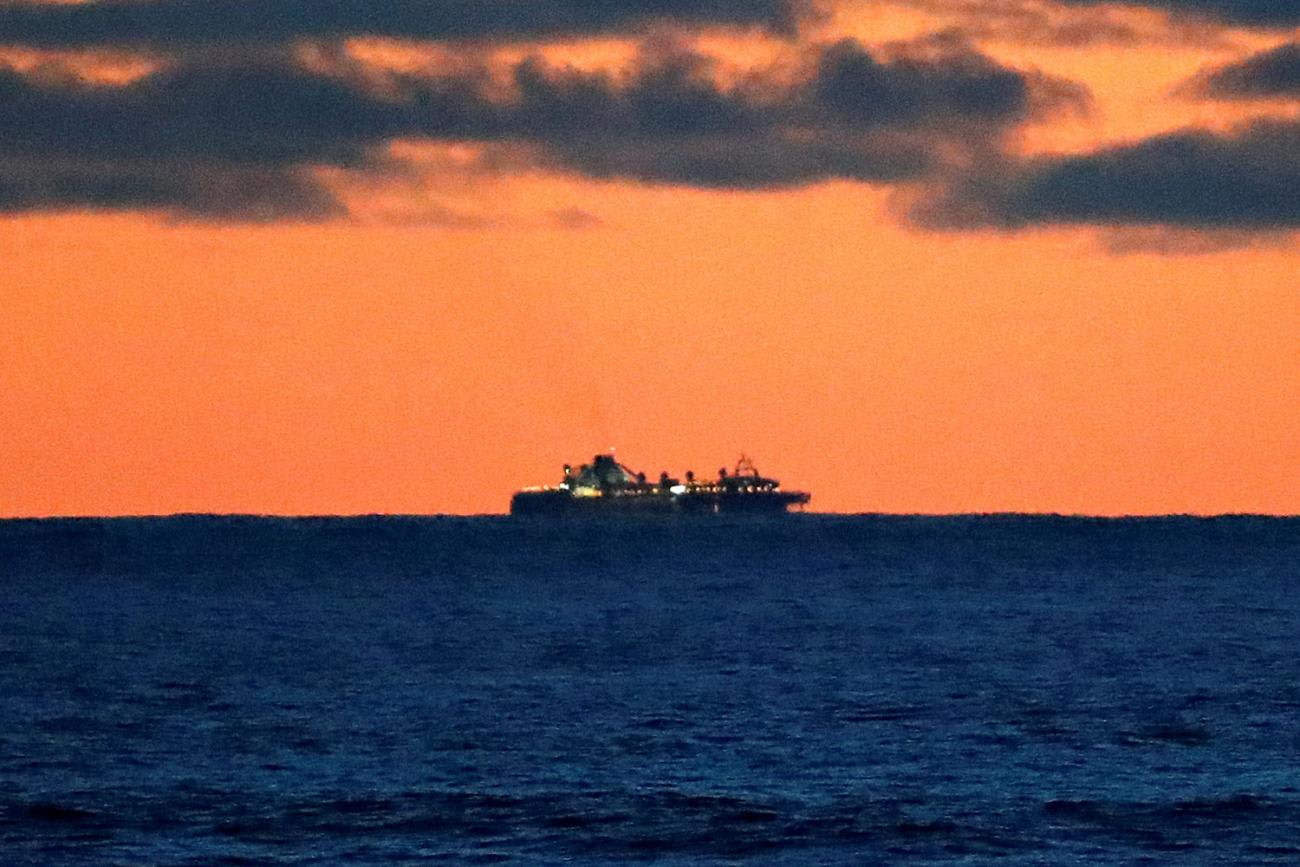 The photo is an open ocean shot of the large cruise ship far off in the distance against a brilliant sunset.
