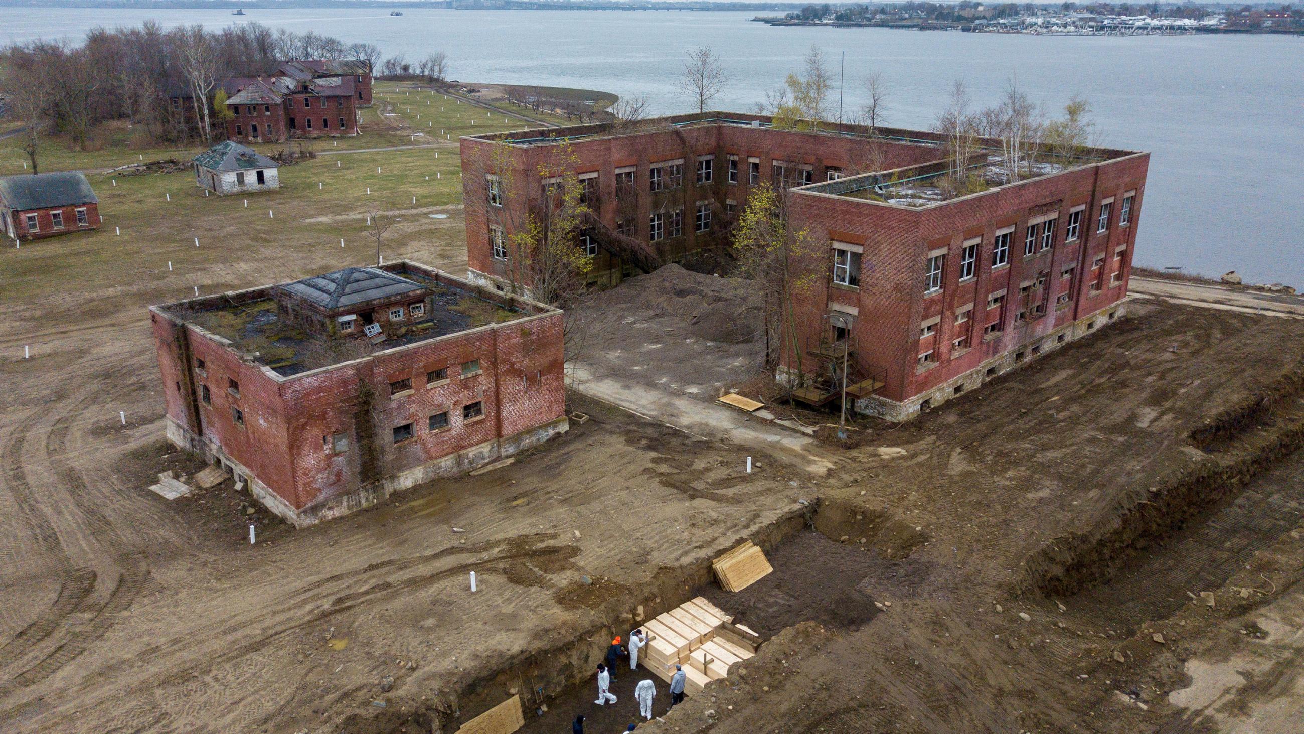 The image shows a long, deep trench into which pine box caskets are being stacked three high and two deep.