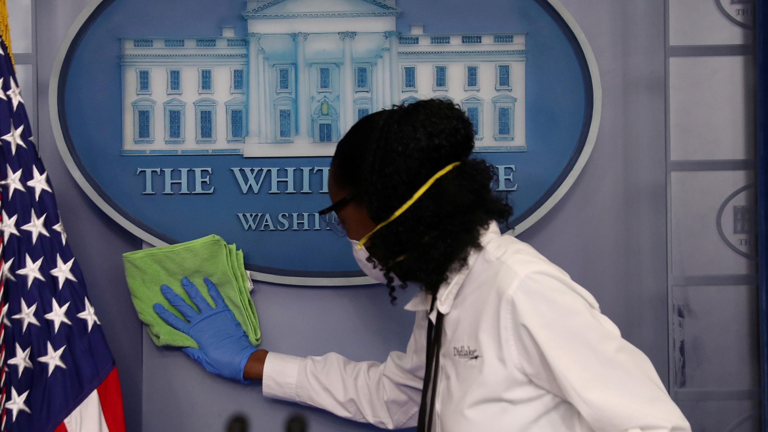 This is a striking image for its subject matter. It's a shot of the podium we don't normally see—empty of politicians and instead a worker in protective gear is cleaning the White House emblem.