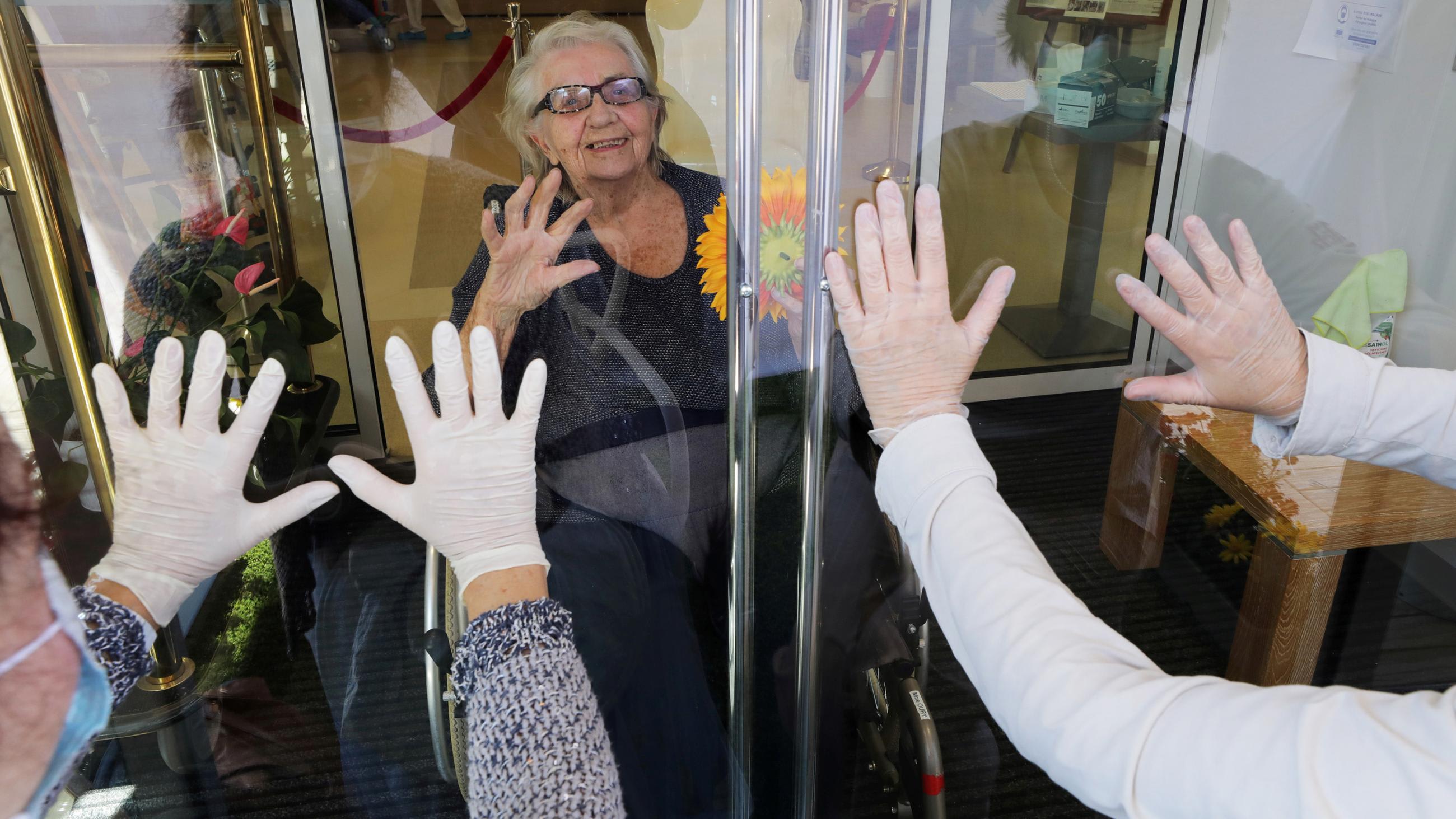 The photo shows an elderly lady looking out at delight while her daughters' hands are held up to the glass.