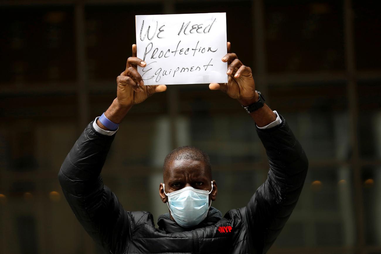 The image shows a man wearing a mask and holding a small sign above his head that says, "We need protection equipment."