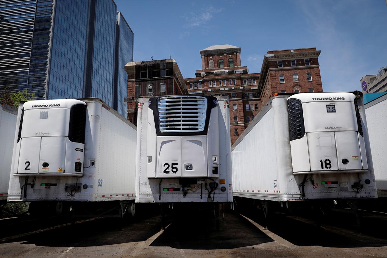 Image shows a row of huge refrigerated trucks parked together amid buildings under a clear blue sky.