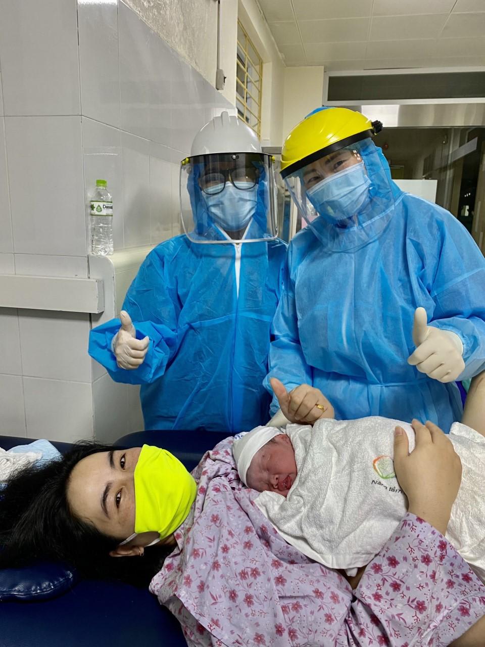 A woman in a bright yellow mask smiles as she holds her newborn infant to her chest. Behind her, two medical professionals in blue medical smocks and masks give a thumbs up and smile.