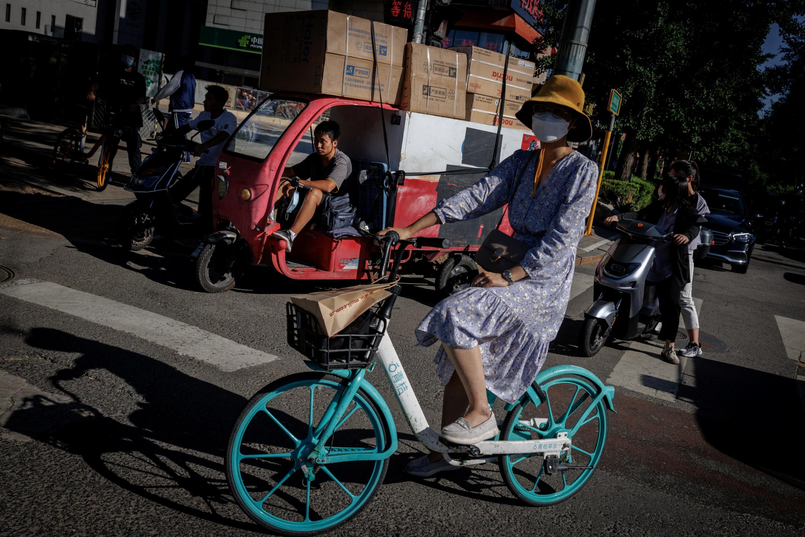 A woman on an aqua-colored bike in a dress and sneakers waits with people at a traffic light in Beijing, China, on July 14, 2022.