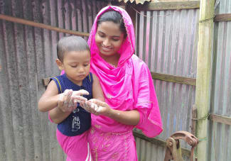 Health leader Sabikunnahar, wearing a bright pink traditional outfit, smiles while helping a young child wash their hands with soap and water outdoors beside a corrugated tin wall.